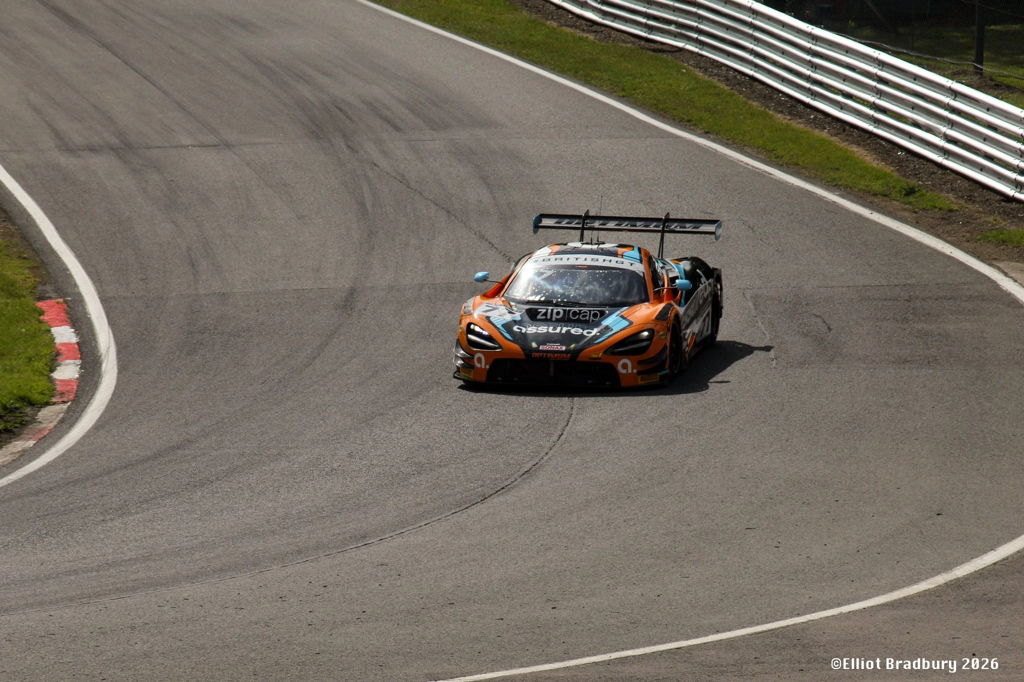 A race car on a curving race track, with guardrails and grass on the sides.