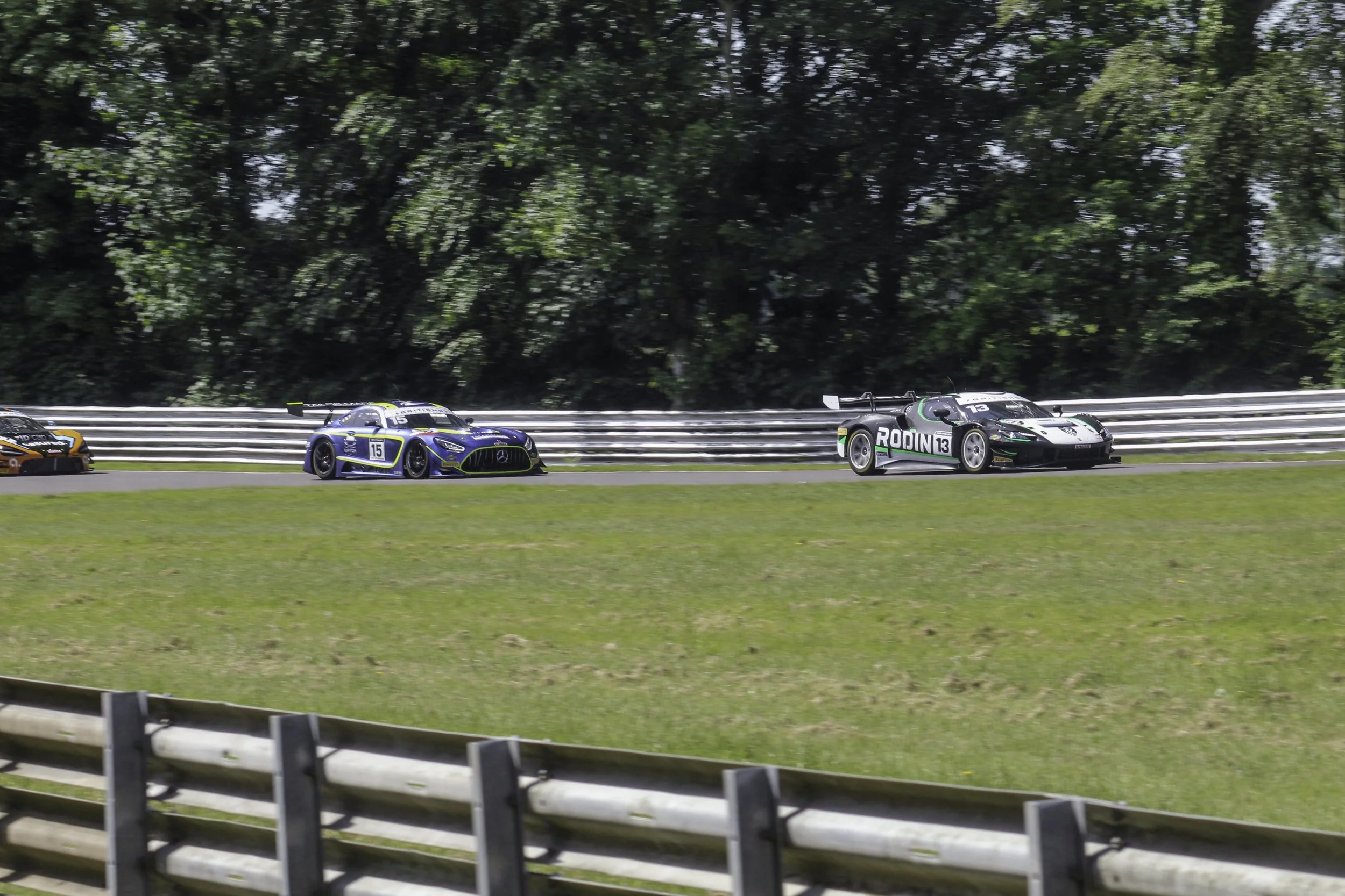 Race cars speeding on a track with green trees in the background and a metal guardrail in the foreground.