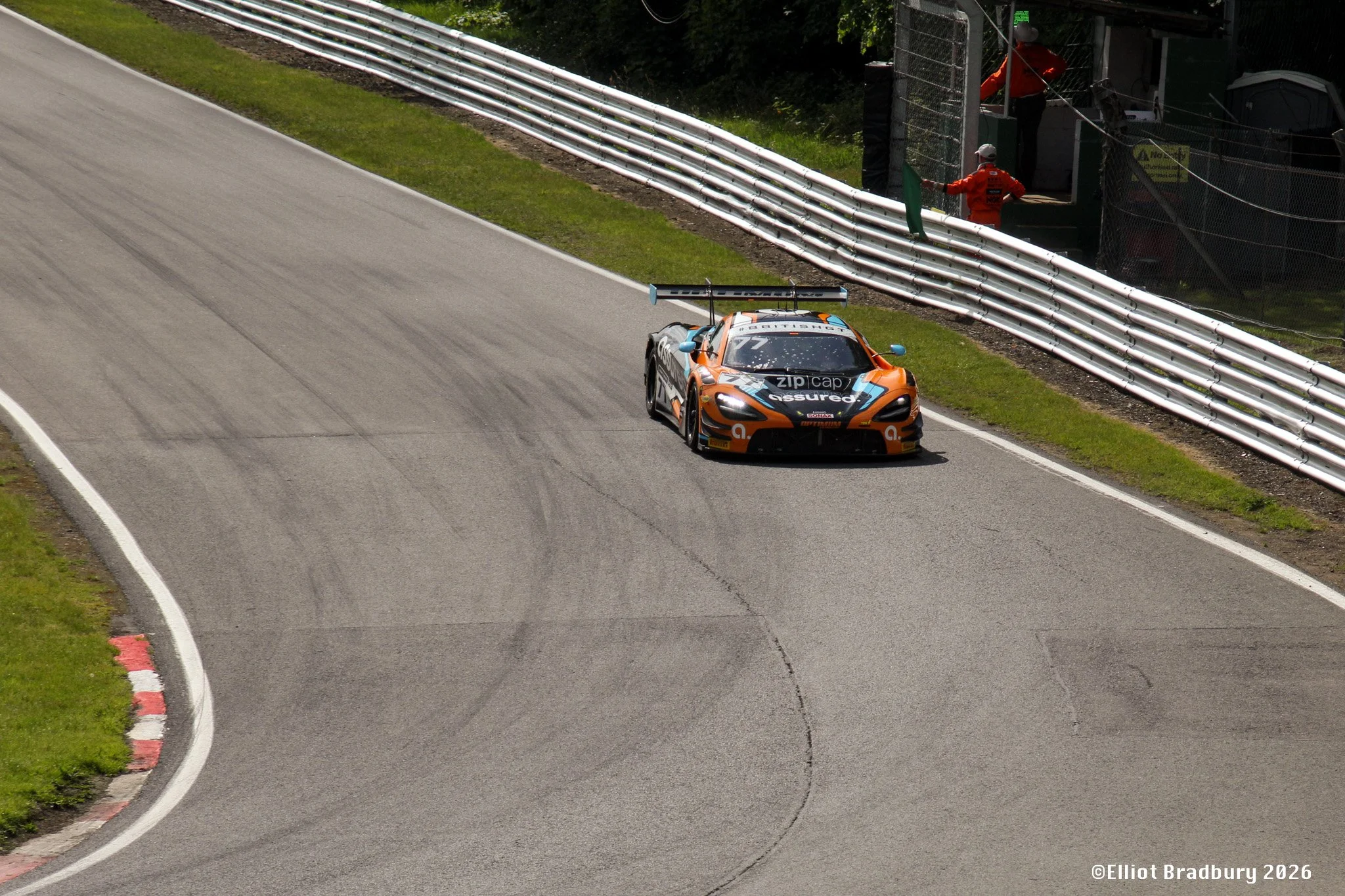 A race car on a racetrack near the pit lane, with two race officials or crew members standing on the sidelines behind safety fences, one in a red jumpsuit and one in black. The car is orange and black, with a prominent rear wing, and has sponsor deca