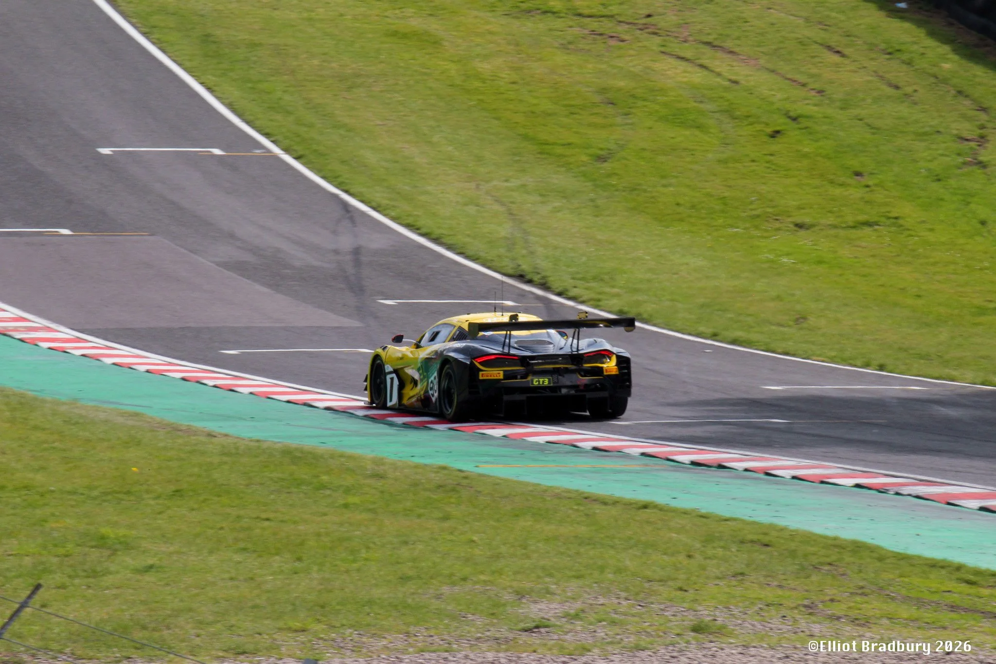 A yellow and black race car on a race track, approaching a curving section with green painted run-off area and grass on the sides. The car has the number 7 on its side and a large rear wing.