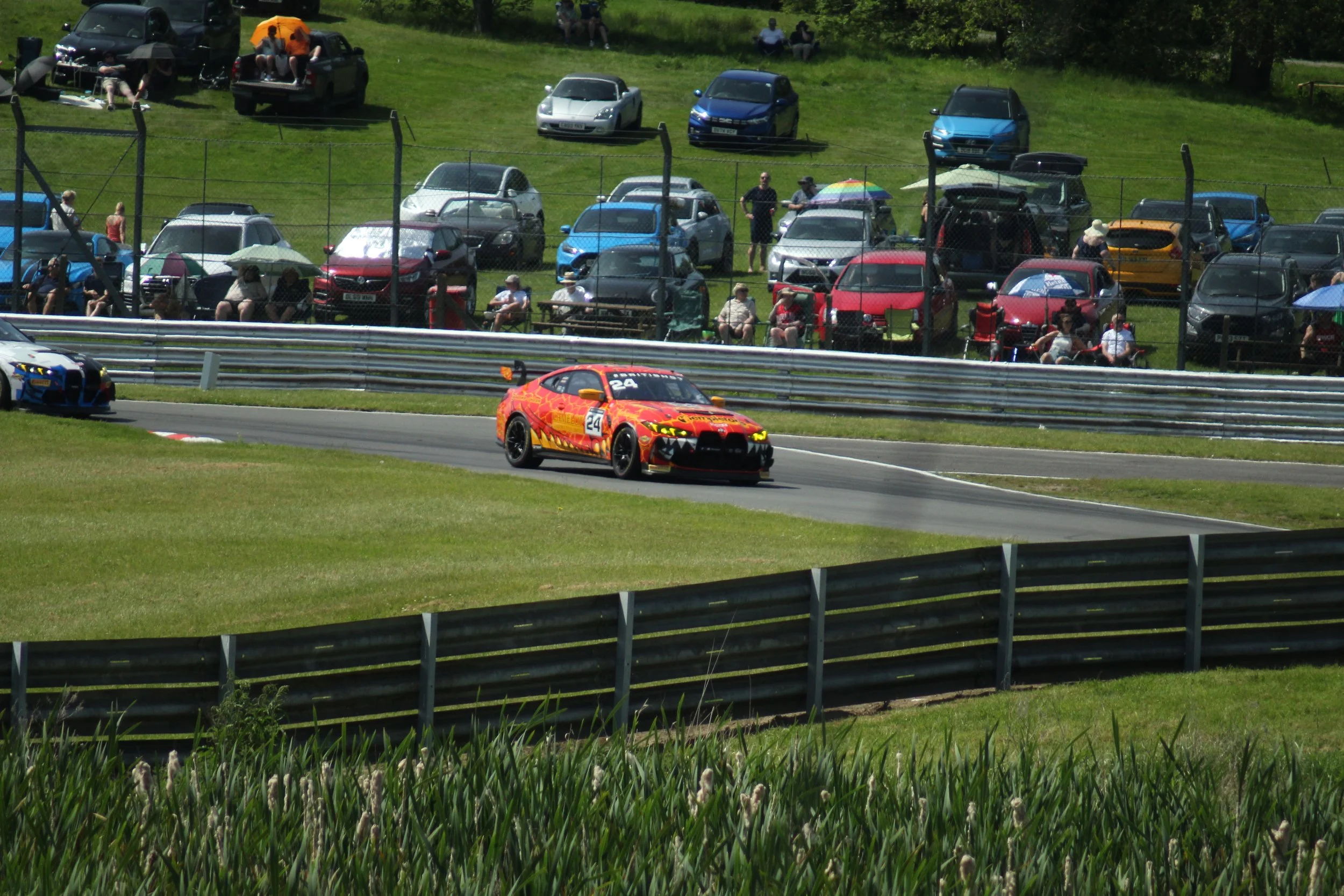 Race car on a race track with spectators watching from the grassy hillside behind a fence.