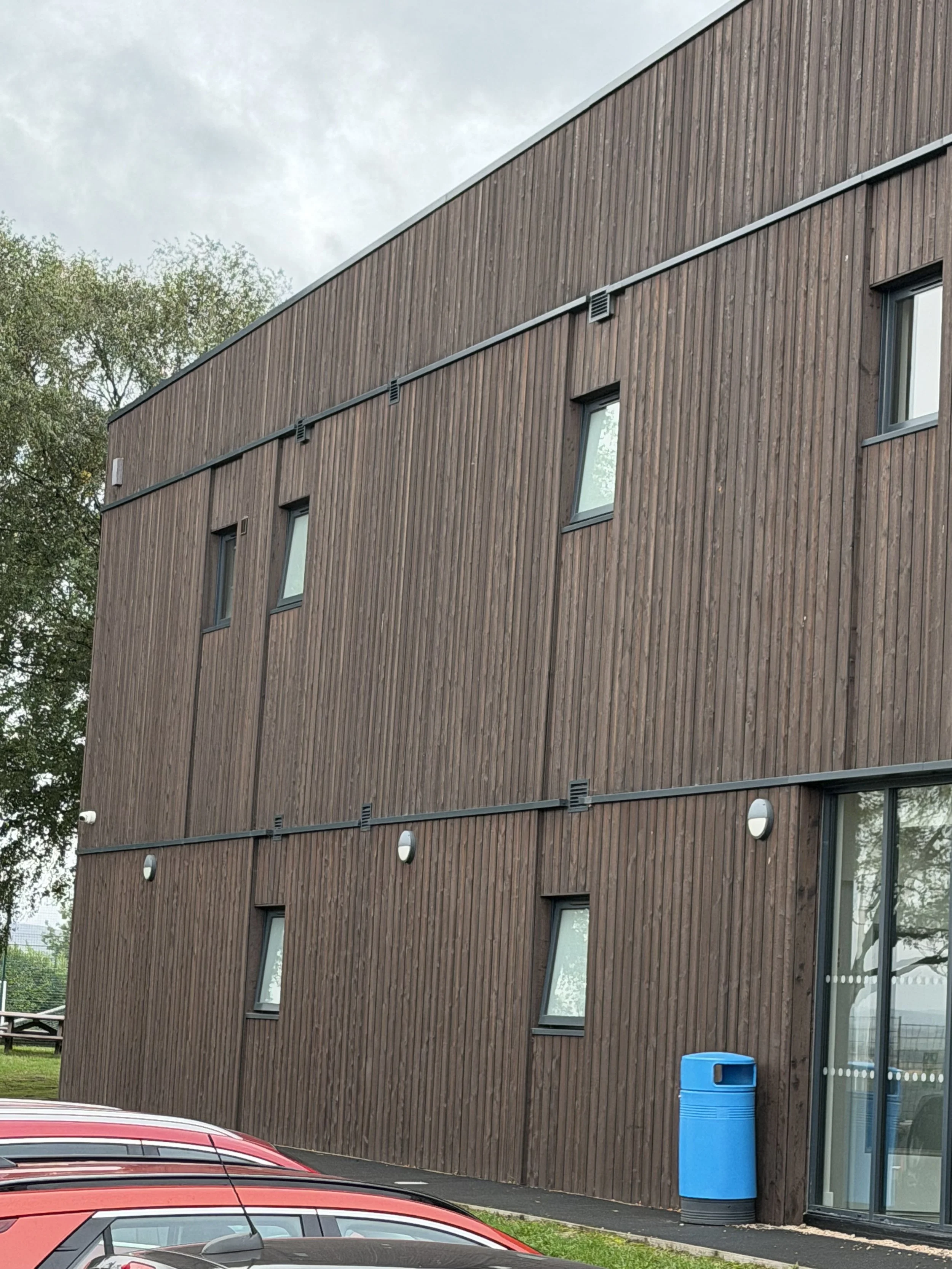 A modern building with wooden siding, multiple small windows, a blue trash bin outside, and parked cars nearby, overcast sky in the background.