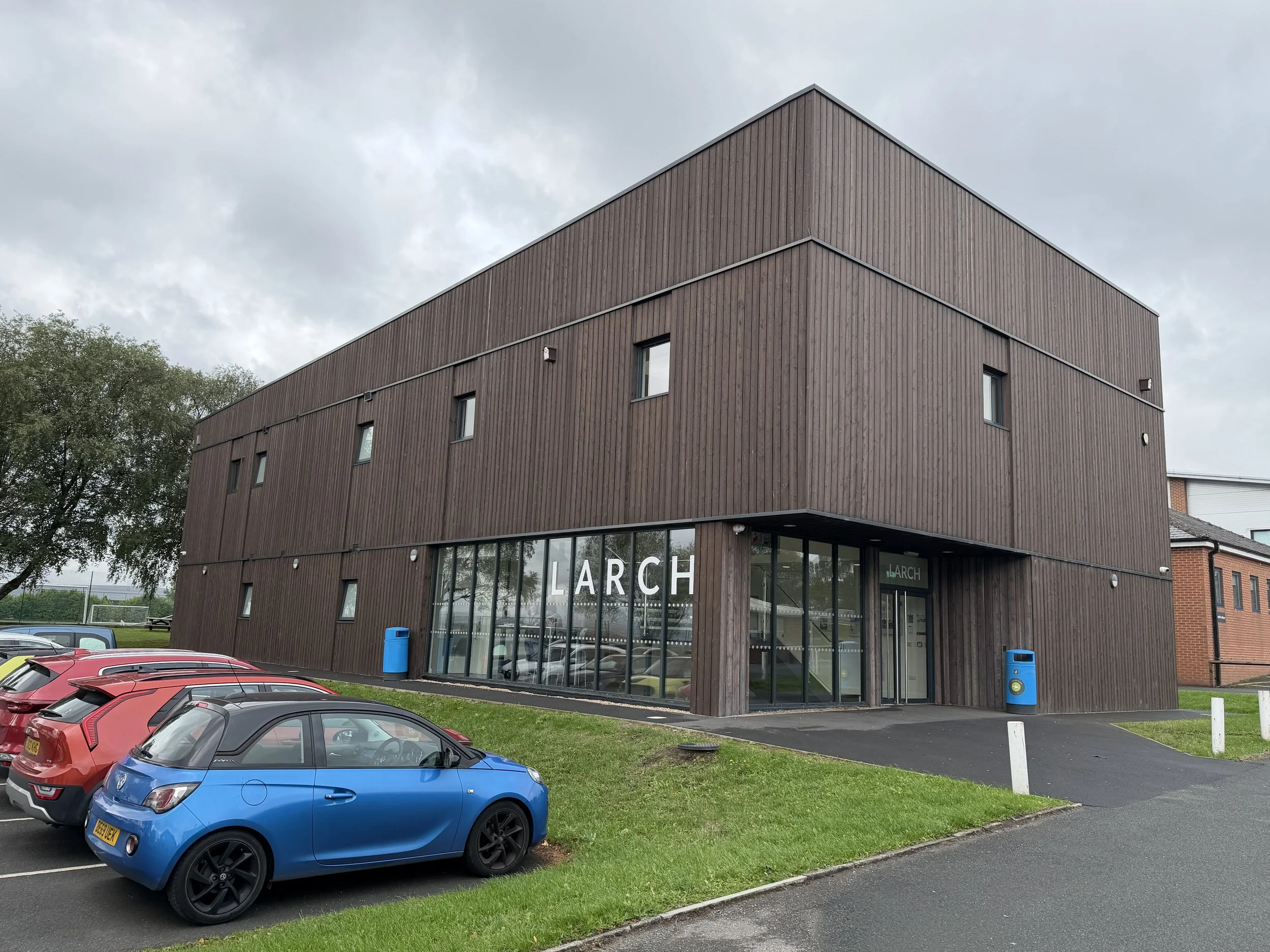 A modern, three-story building with a wooden exterior and large glass storefront on the ground floor. There are several small, rectangular windows on the upper floors. The building has the word 'LARCH' on the storefront. In front, there are parked ca