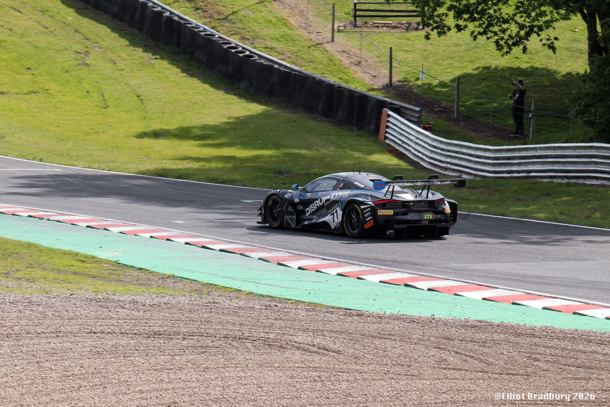 A race car on a racetrack with green striped curbing, a grassy hillside, a black tire barrier, and a spectator standing behind a fence taking photos.