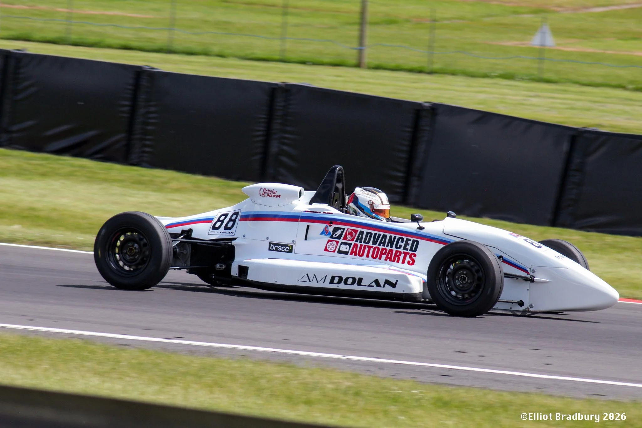 A white race car with multiple sponsor stickers, including ADVANCED AUTOPARTS, AM DOLAN, and others, racing on a track with green grass and fencing in the background.