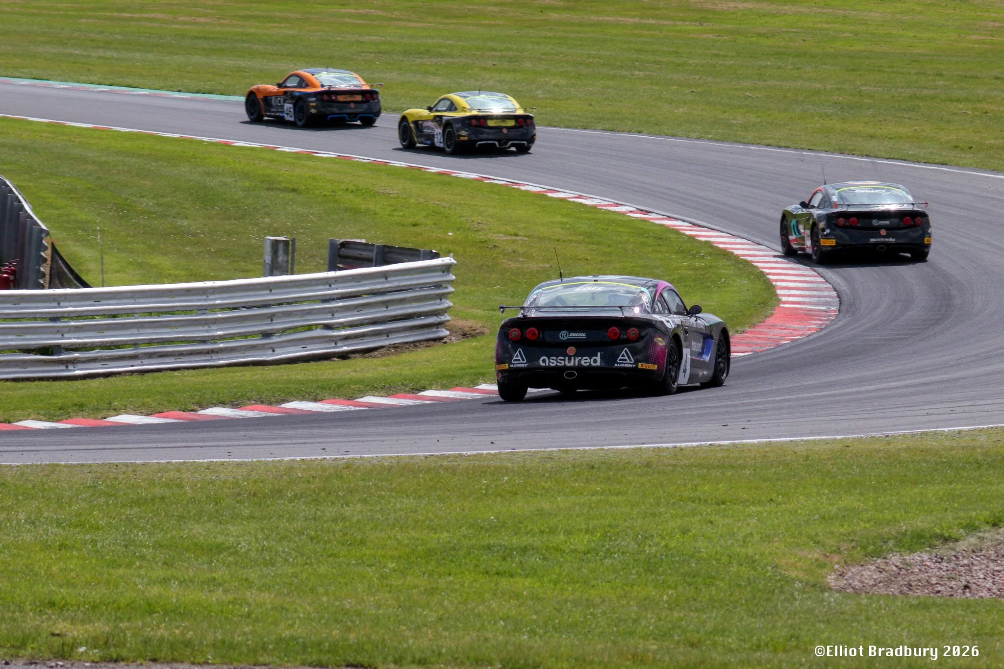 Multiple race cars on a race track, navigating a curve with grass on either side, safety barriers on the left.
