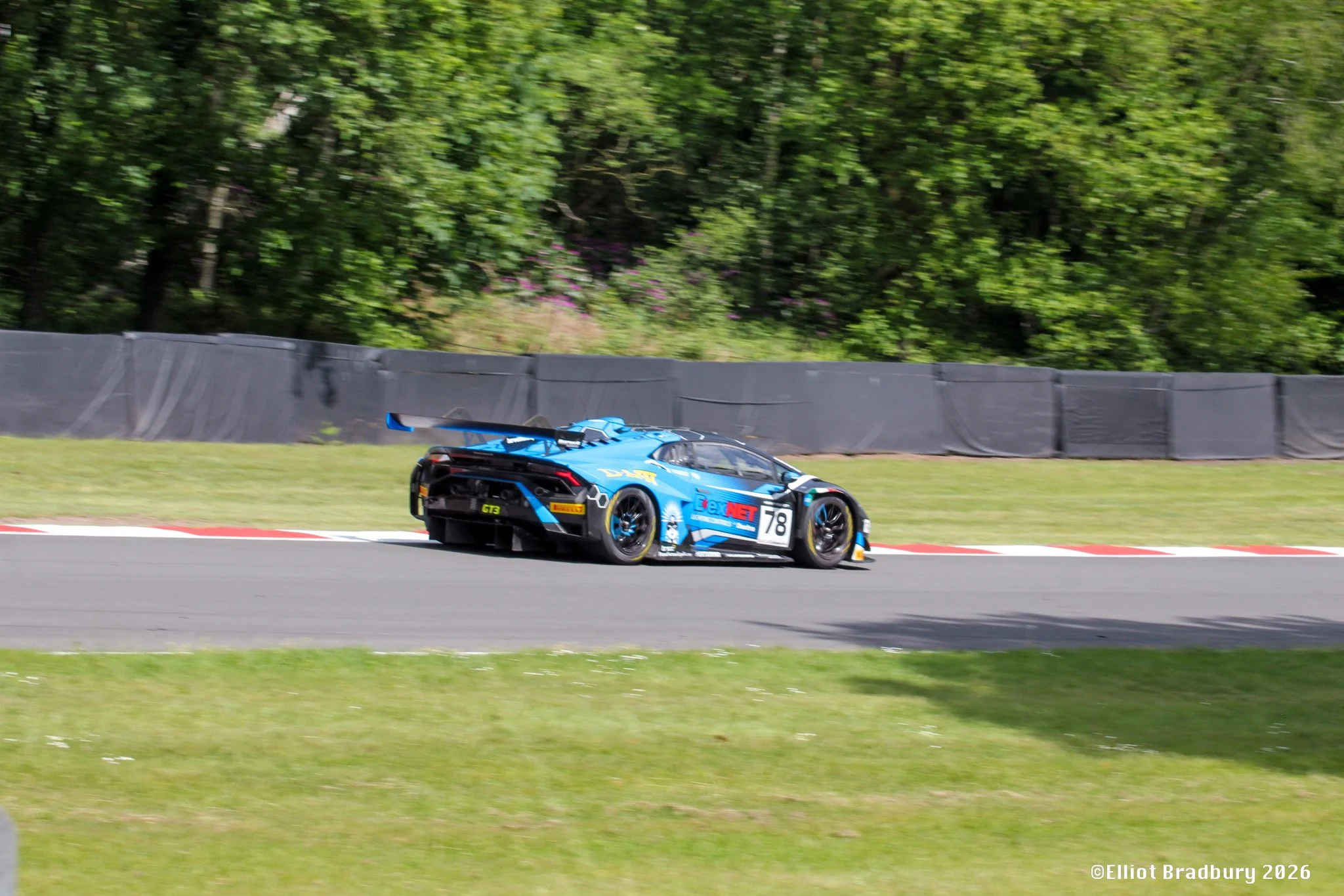 A blue race car on a track, with a forested background and a black barrier