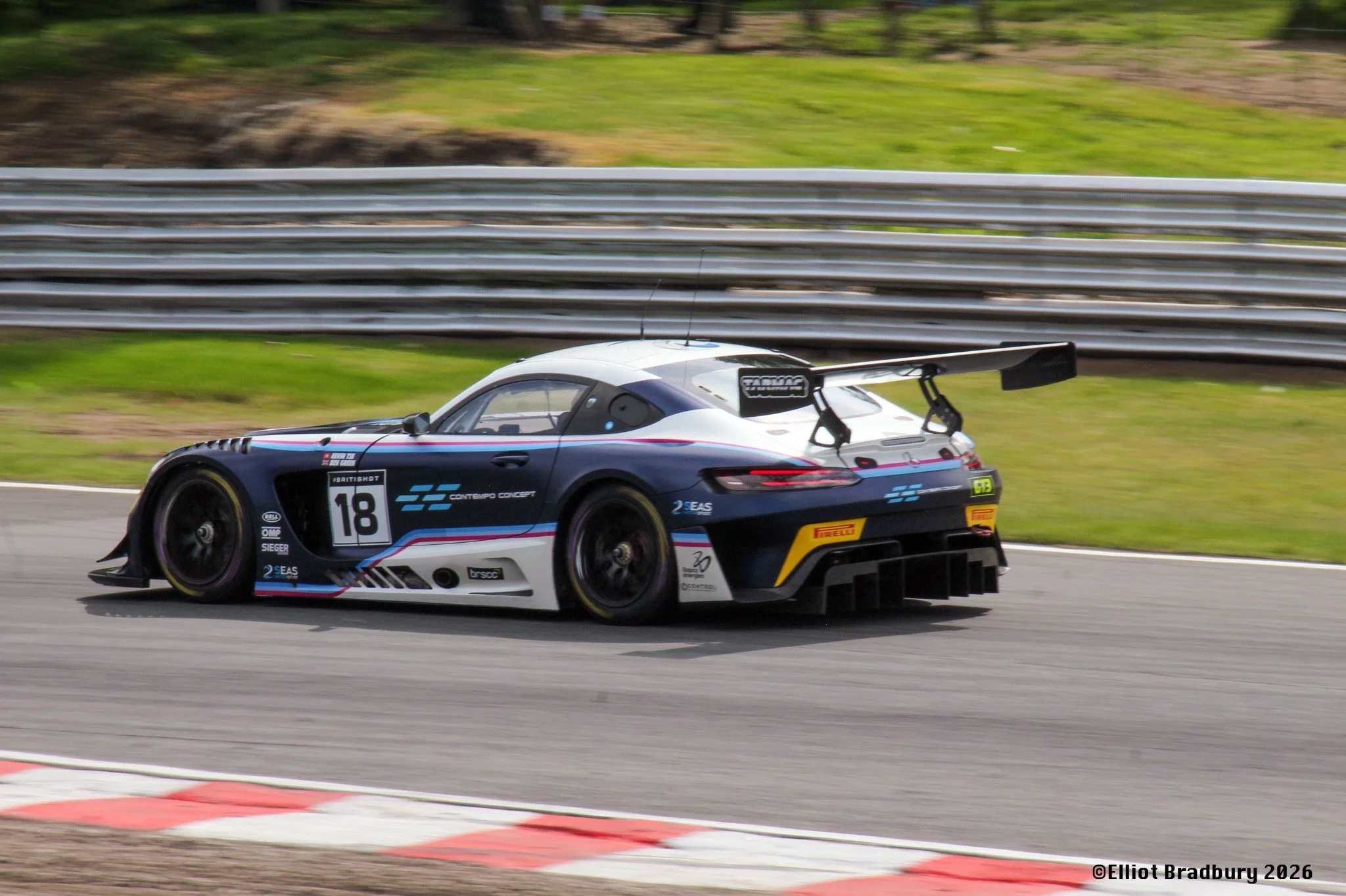 A race car with black, white, and blue livery on a racetrack, passing a red and white striped curbing, with a safety barrier and greenery in the background.