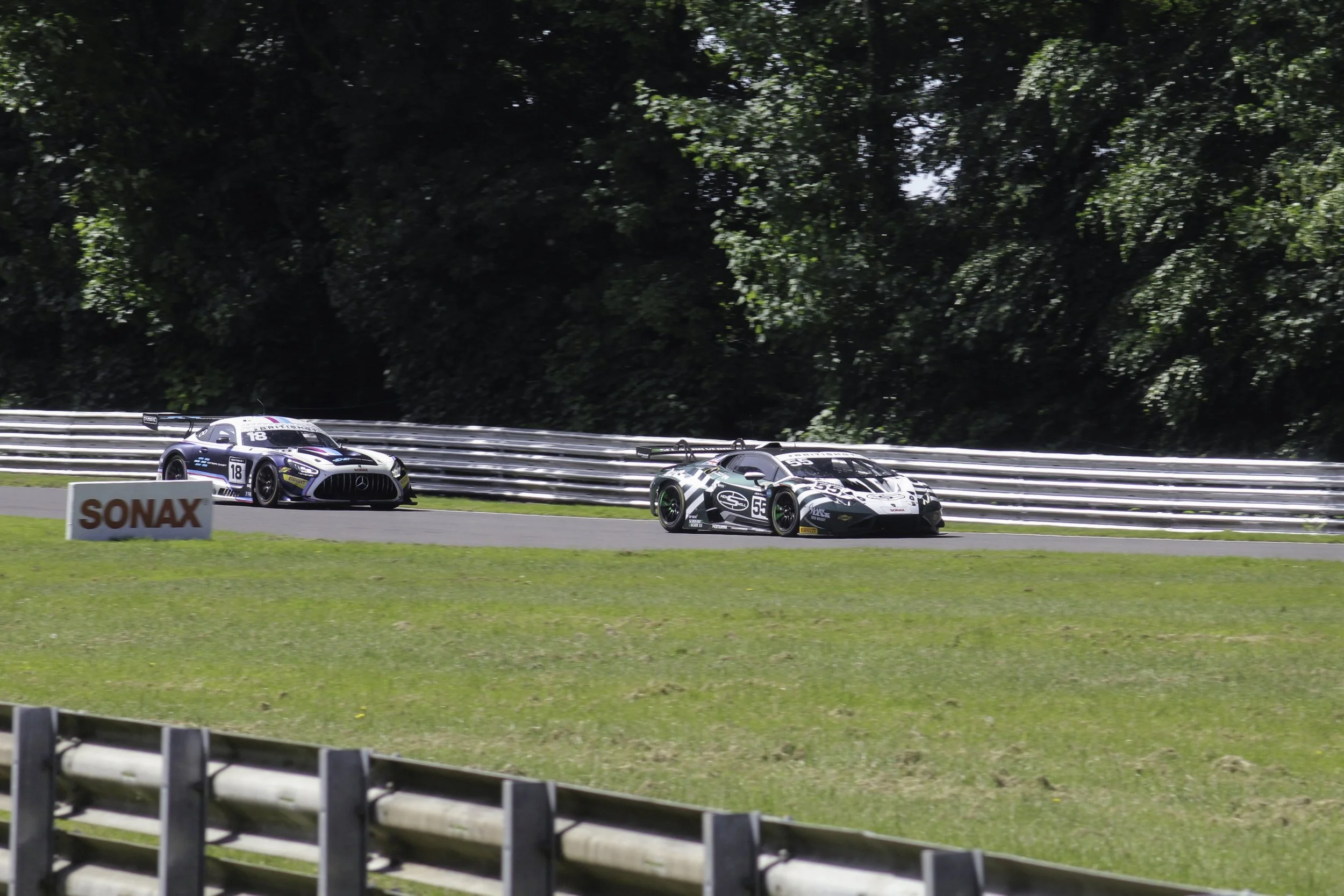 Two race cars speeding on a track, with a backdrop of lush green trees and a metal safety barrier, and a sign reading 'SONAX' in the foreground.