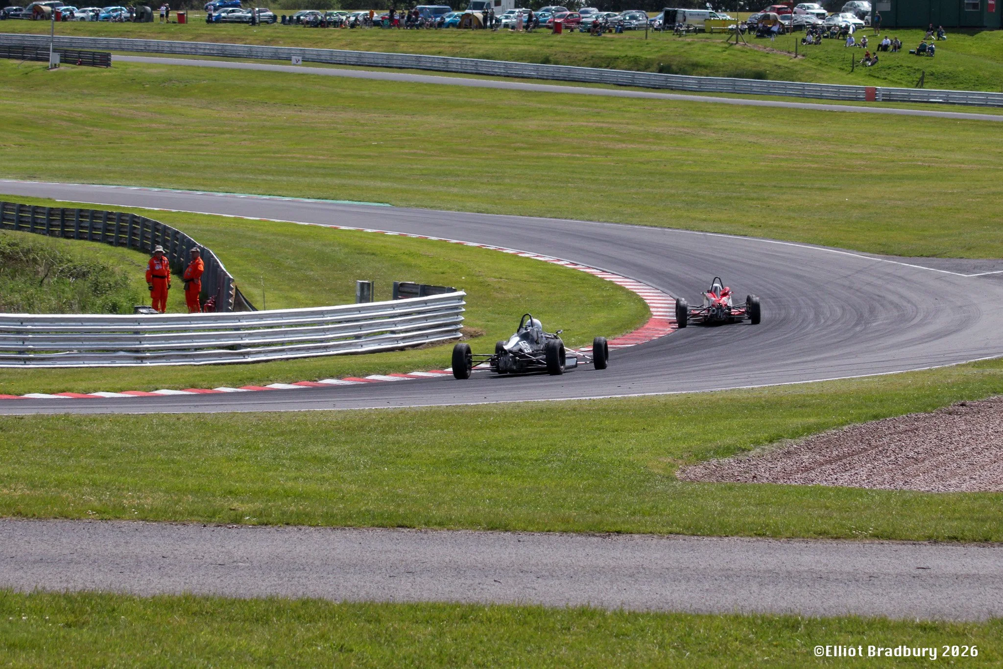 Race cars on a racetrack with grass and spectators in the background.
