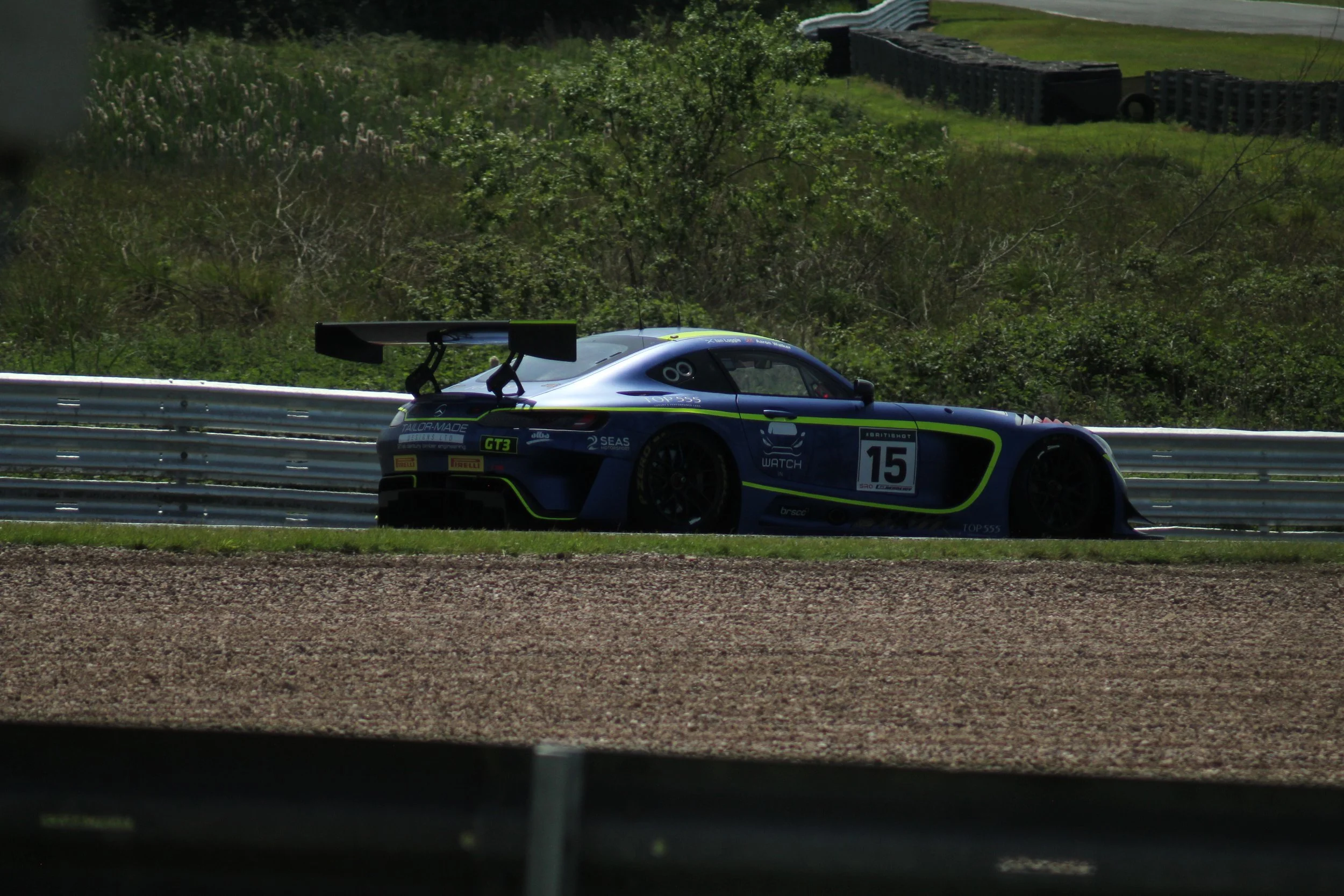 A blue race car with a large rear spoiler driving on a racetrack, seen from inside a vehicle or behind a barrier with greenery and tires alongside the track.