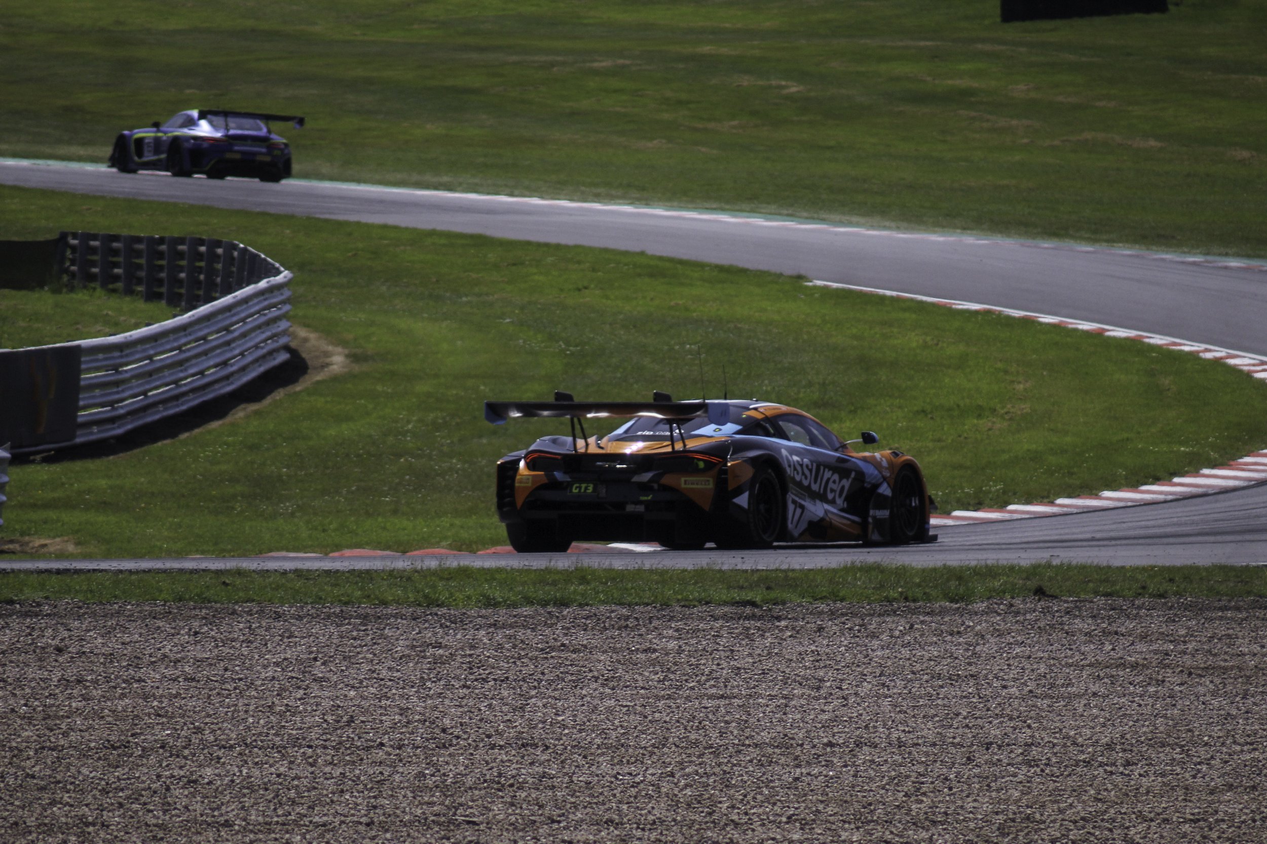 Two race cars on a racetrack, one orange and black and the other black with green accents, surrounded by green grass and safety barriers.