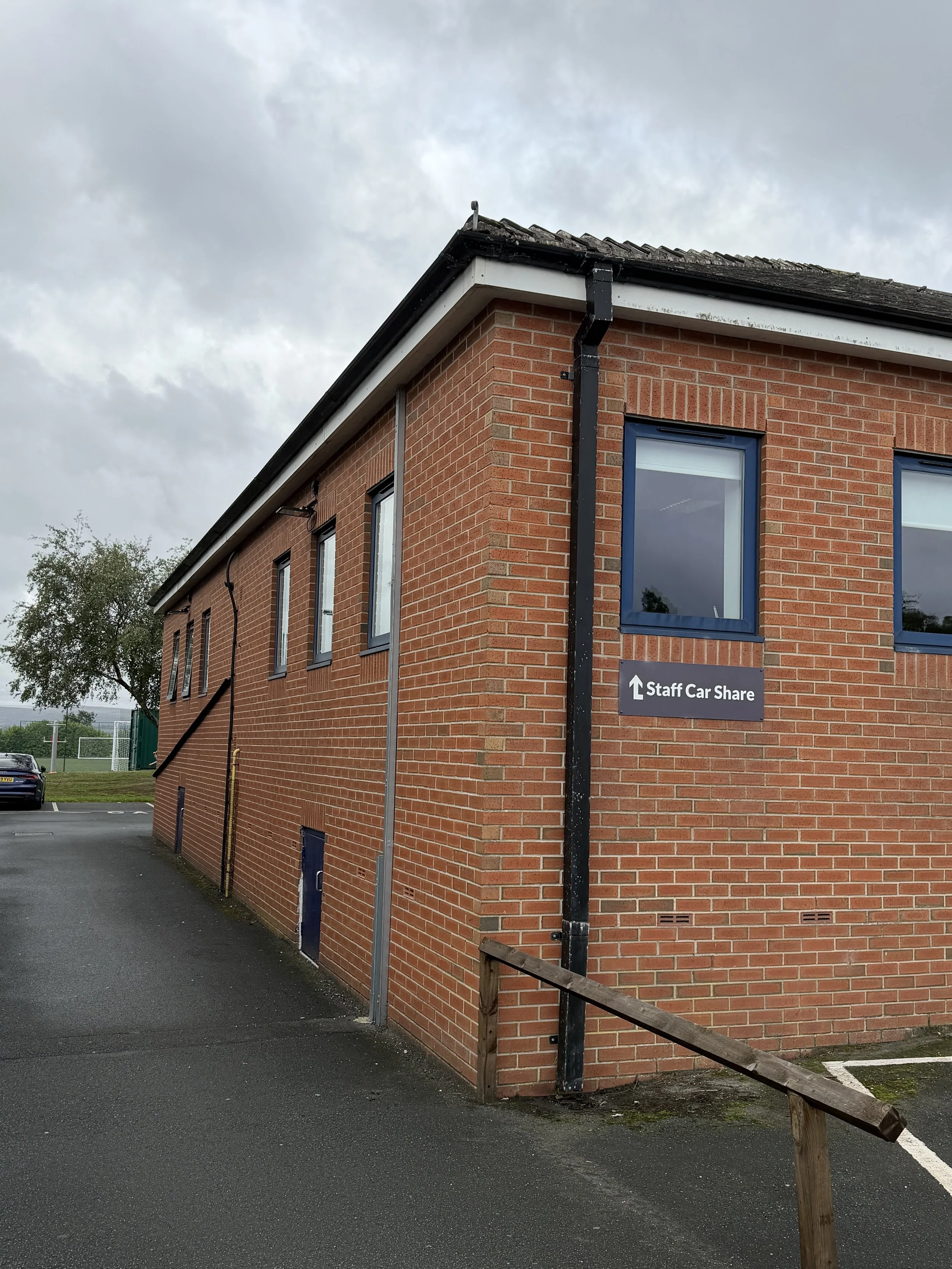 A corner of a brick building with a black gutter and a sign that reads 'Staff Car Share', pointing upwards. There is a wooden railing and some windows on the building, with a cloudy sky above.