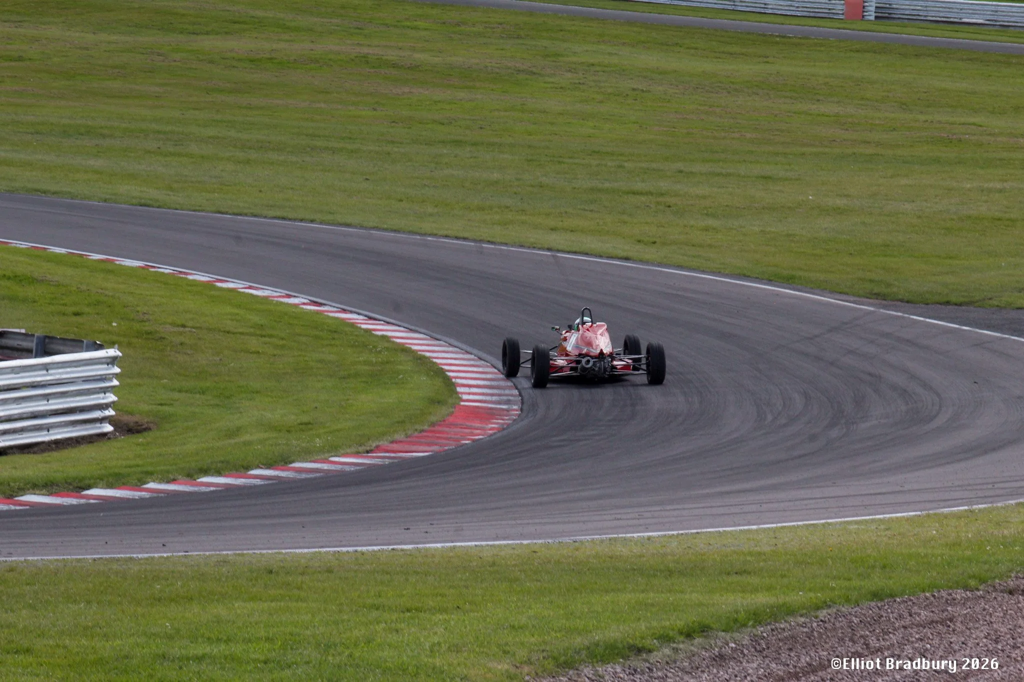 A single small racing car driving on a racetrack, with green grass surrounding the track and cornering markings visible on the surface.