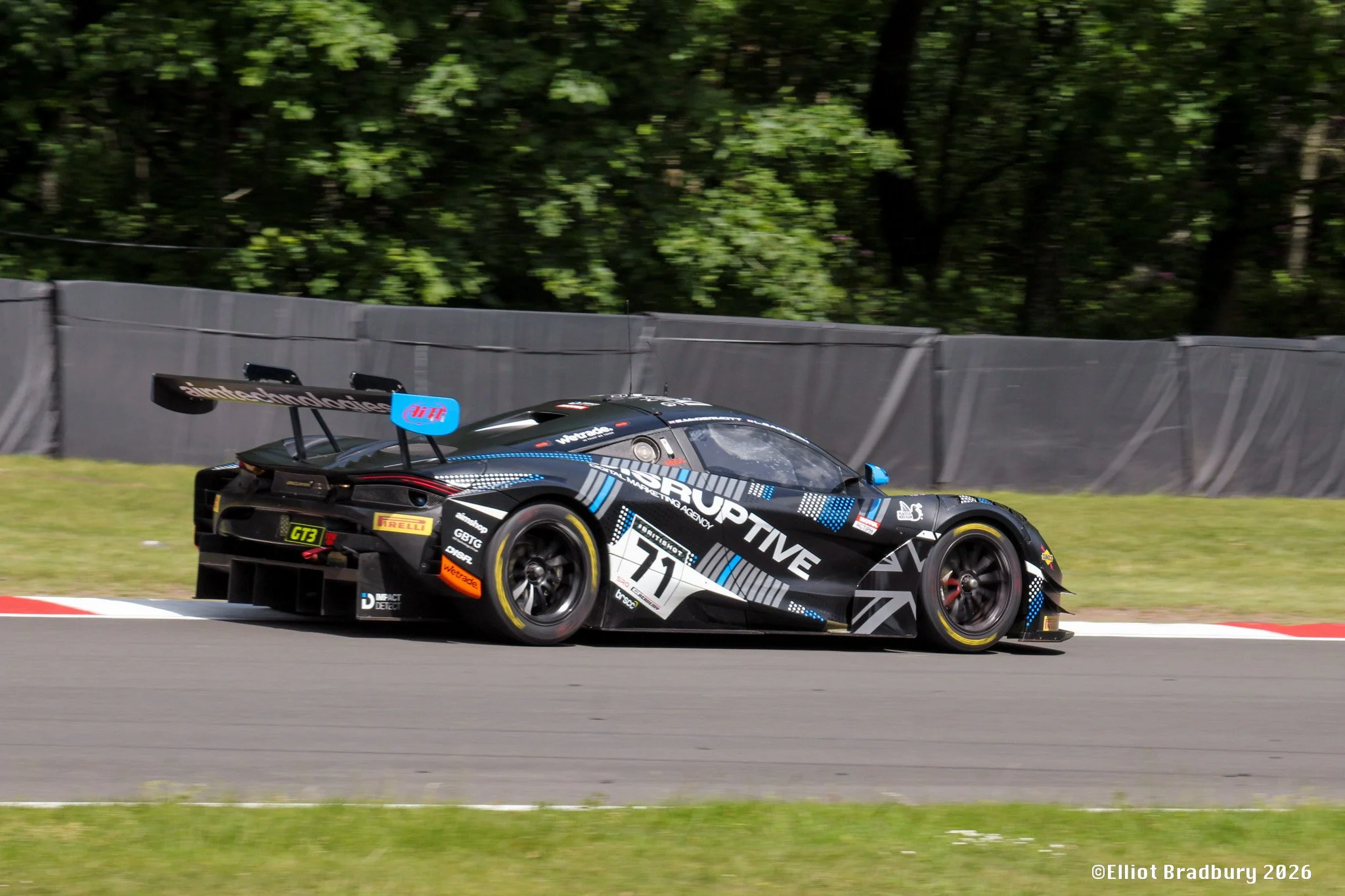 A black race car with white and blue graphics racing on a track, surrounded by green trees and a gray barrier in the background.