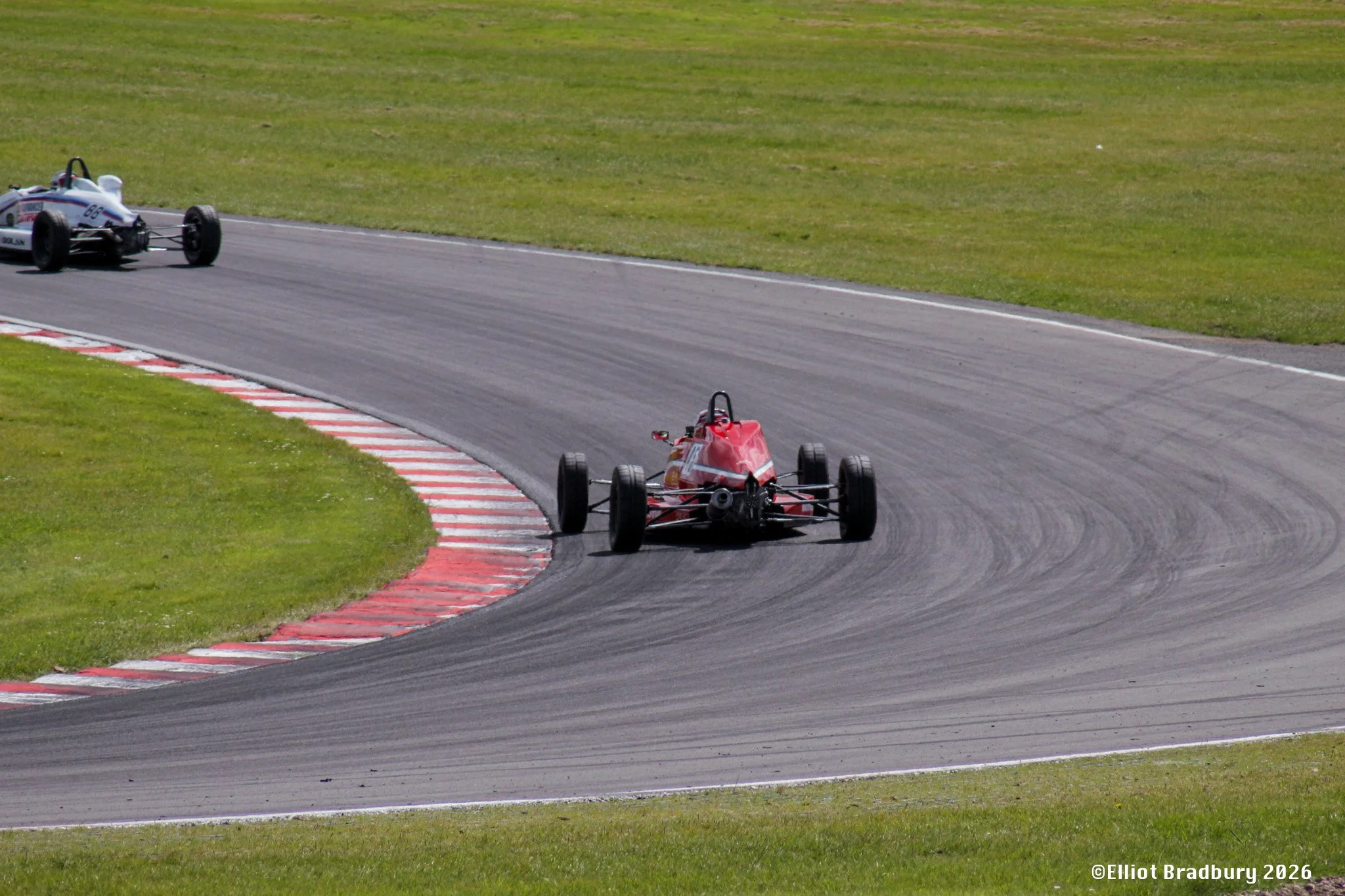 Two race cars on a track, one red and one white with blue, navigating a turn on a grassy racing circuit.