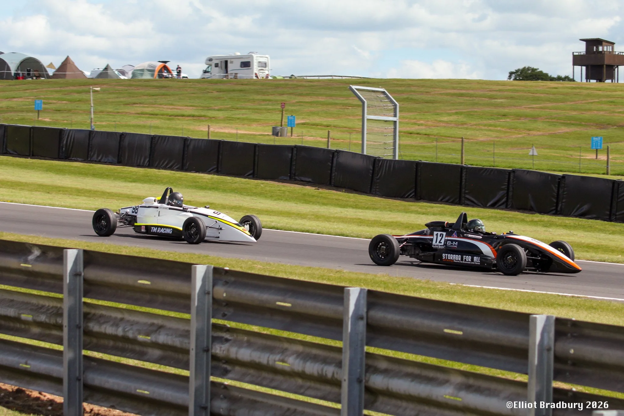 Two race cars driving on a racing track with a black barrier on the side, green grassy hill with tents and a few structures in the background, under a cloudy sky.