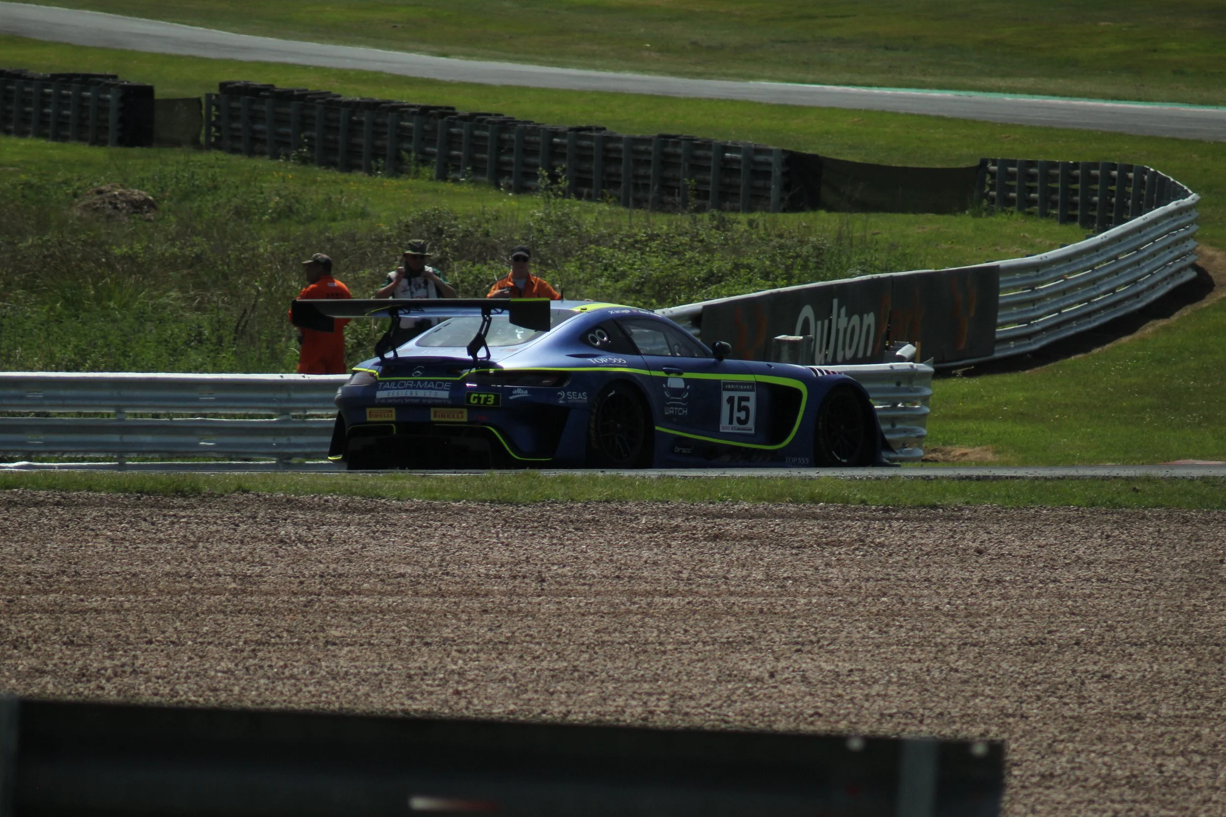 A race car on a track with a crash barrier and three people in orange and white outfits standing nearby.