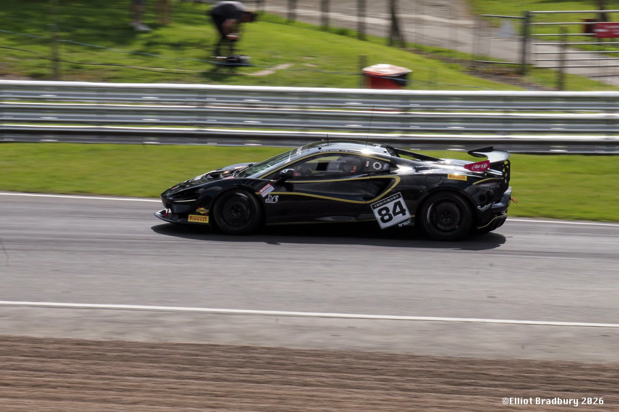 A black racing car with racing stickers and the number 84 on the side, moving on a race track. There is a guardrail in the background and a grassy area with a person in black near a fence further back.