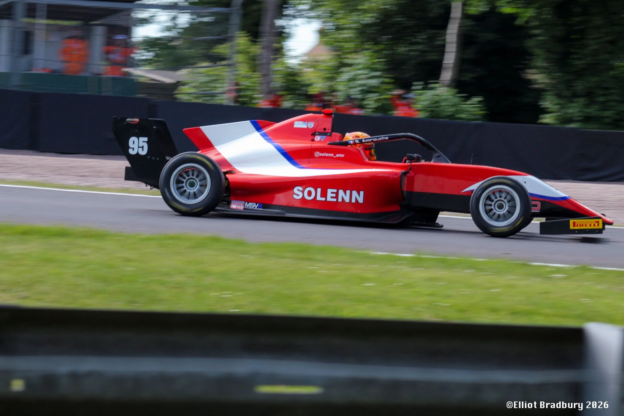 A red and white Solenn racing car with the number 95, speeding on a race track surrounded by green trees and fencing.