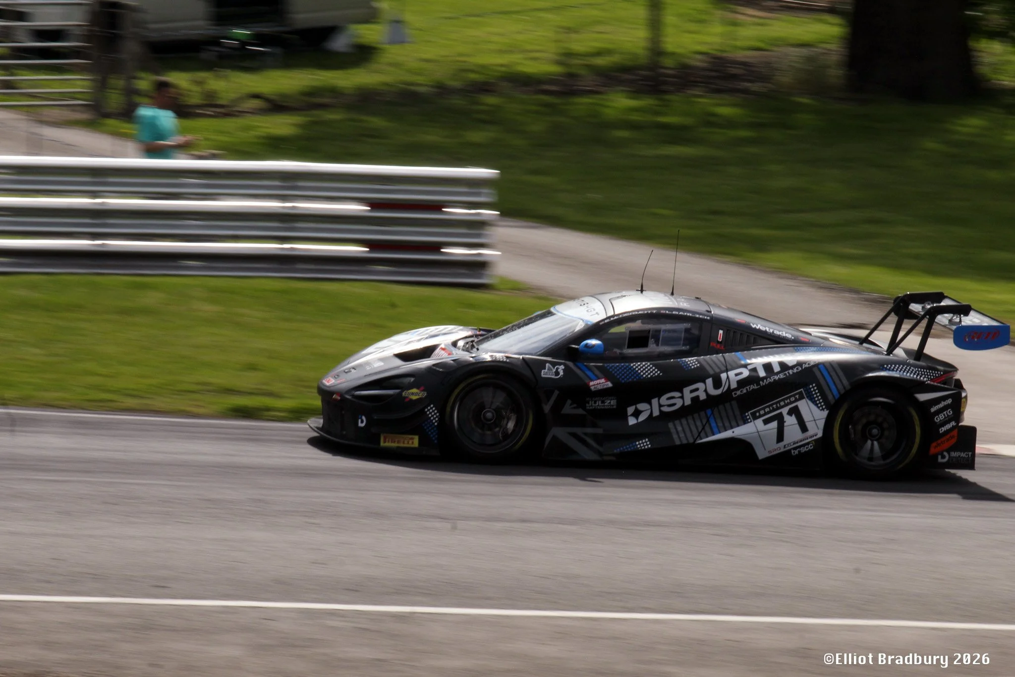 A black and white race car with the number 71 on its side racing on a track, with a spectator behind a metal barrier in the background.