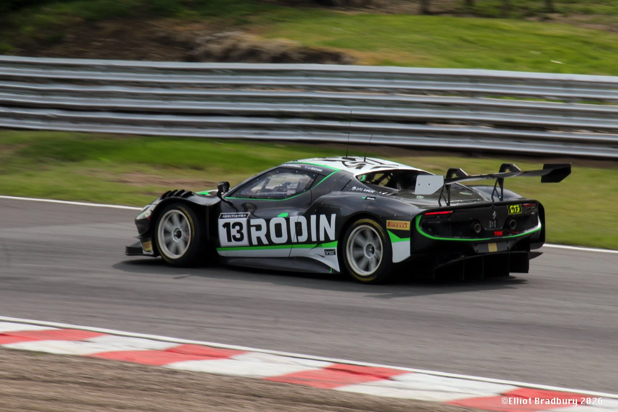 A black and green race car on a racetrack during daytime, with a large rear wing and the number 13 on its side, passing a blurred background of metal guardrails and grassy areas.