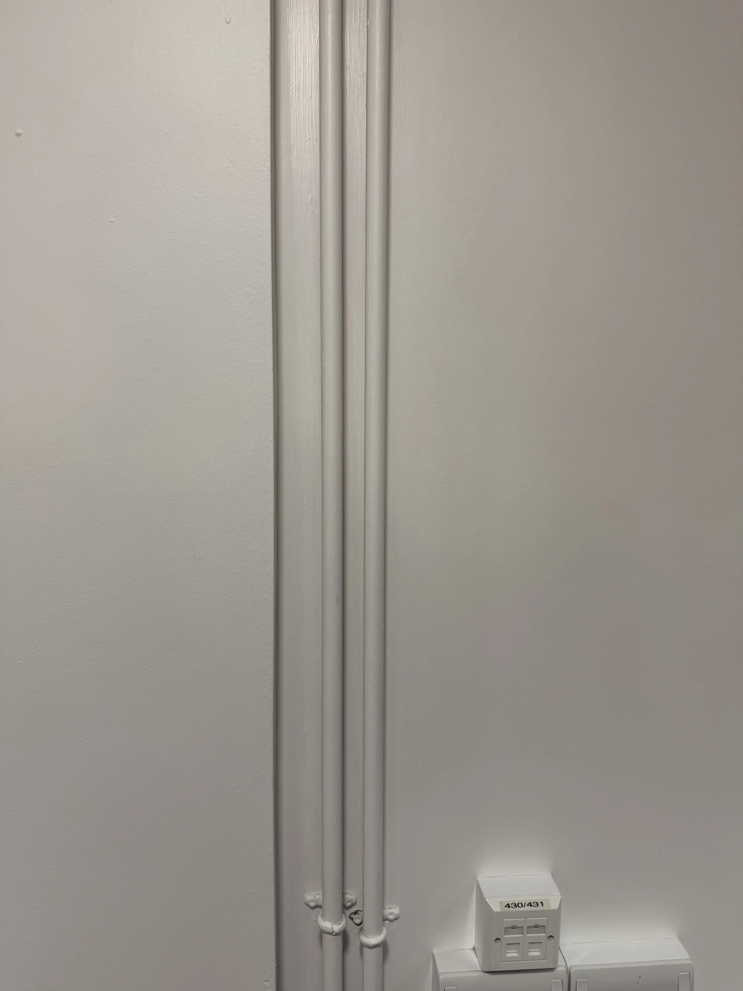 White wall with three vertical painted pipes and electrical outlets below labeled 430/431.