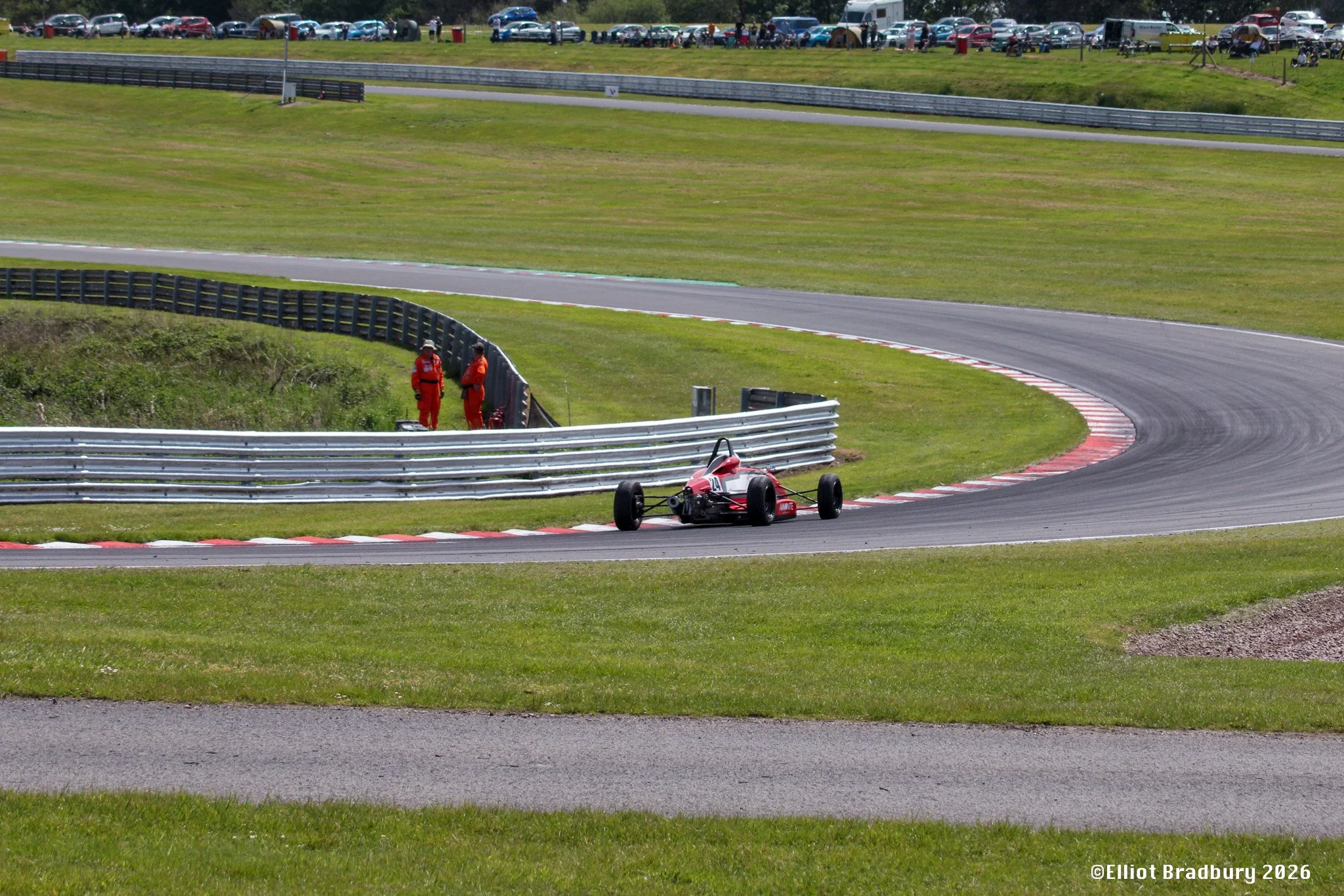 A race track with a red race car approaching a turn, two race officials in red uniforms standing beside the track, and a large parking lot full of cars in the background.