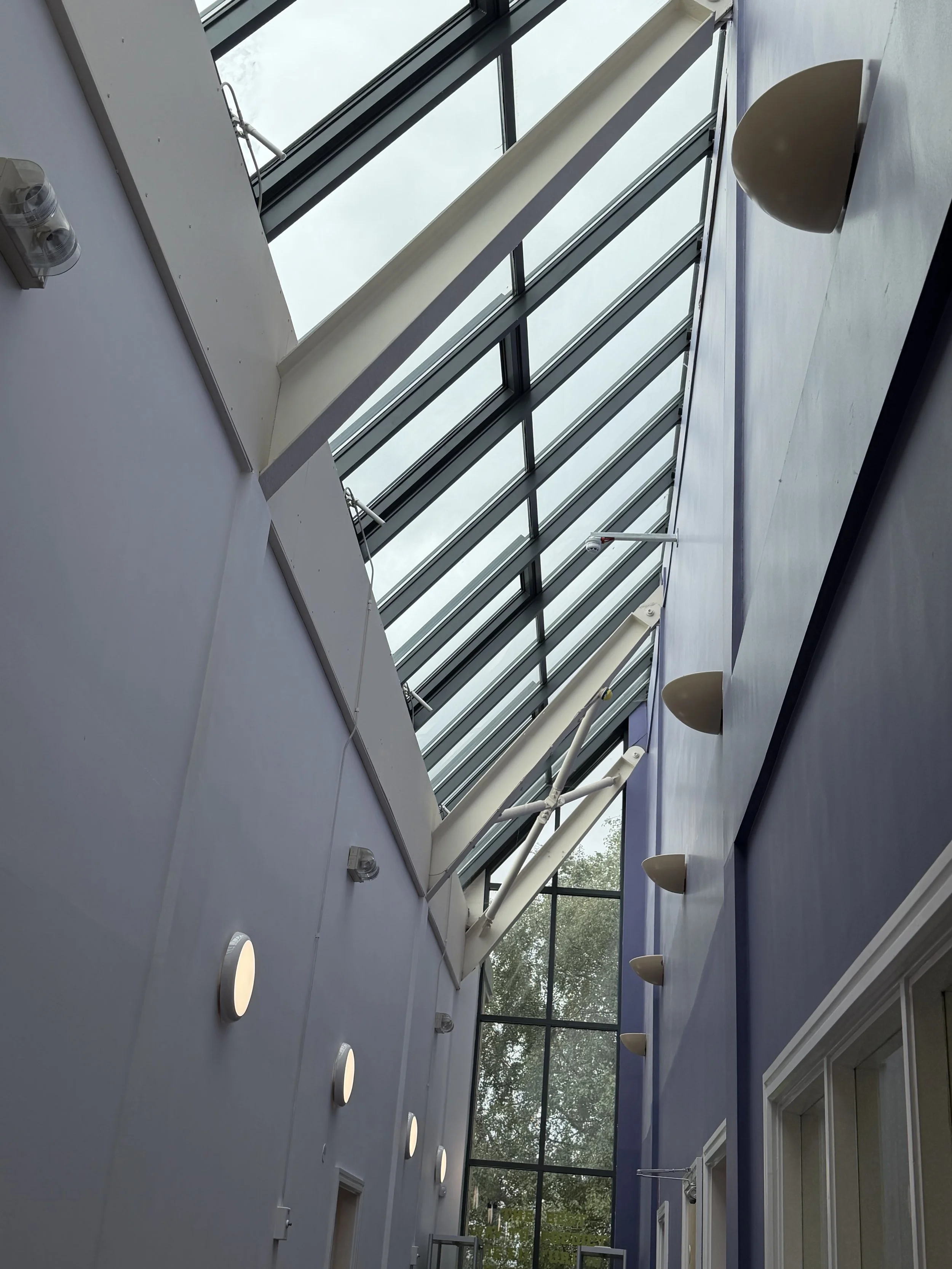 Interior view of a building with large glass windows and skylights, white ceiling with round wall lights, and a blue wall with wall-mounted lights, overlooking outside trees.