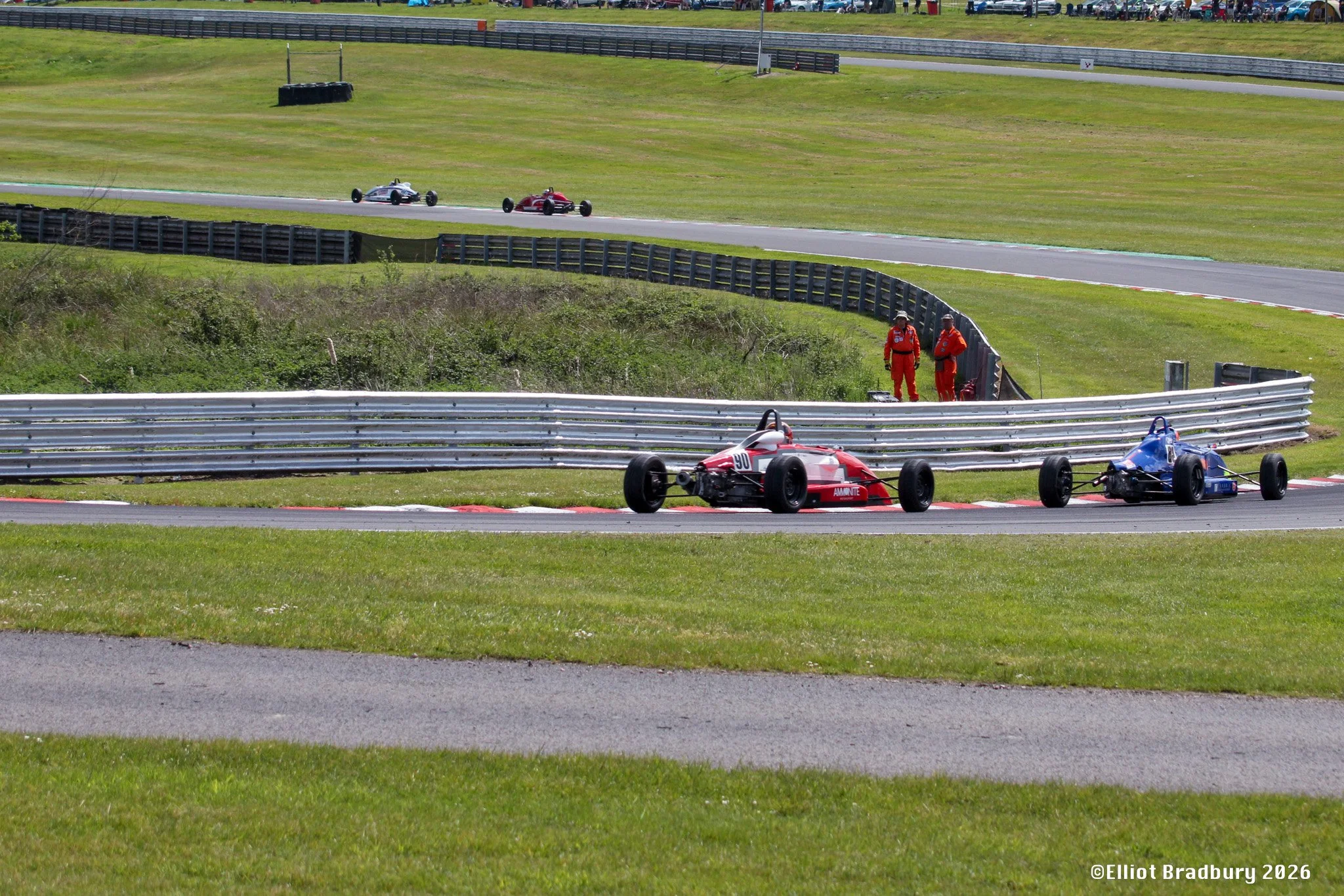 Race cars on a race track with two personnel in red suits near the fence. The cars are small and open-wheel, with one red and one blue, and the track has safety barriers and grassy surroundings.