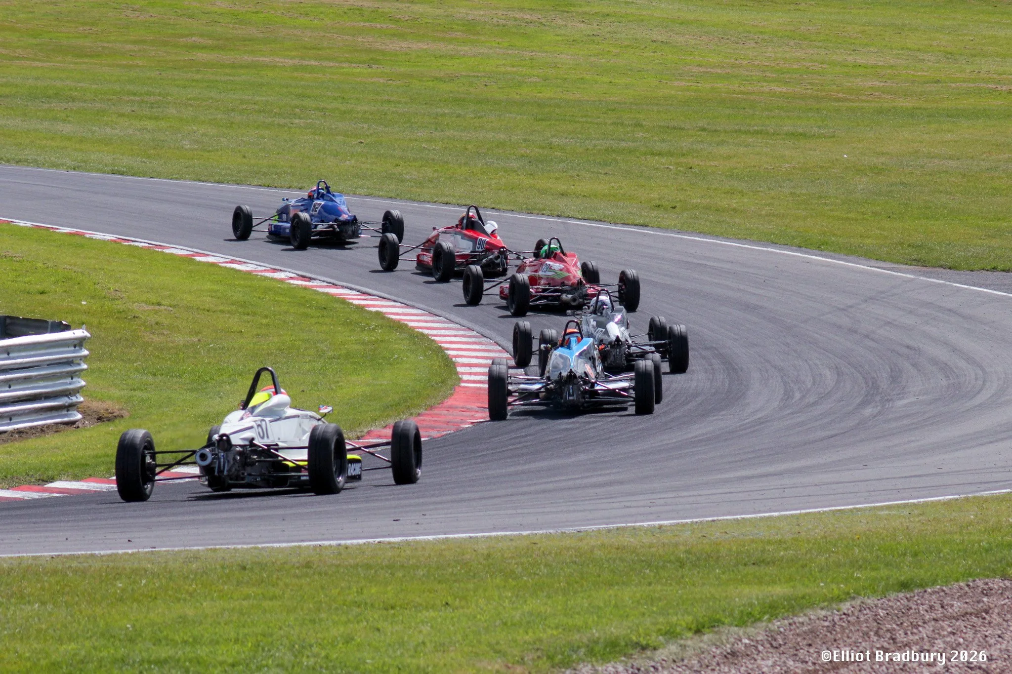 Race cars on a winding dirt track with grassy surroundings, some cars in motion and close to the camera, others further back. Bright and sunny day.
