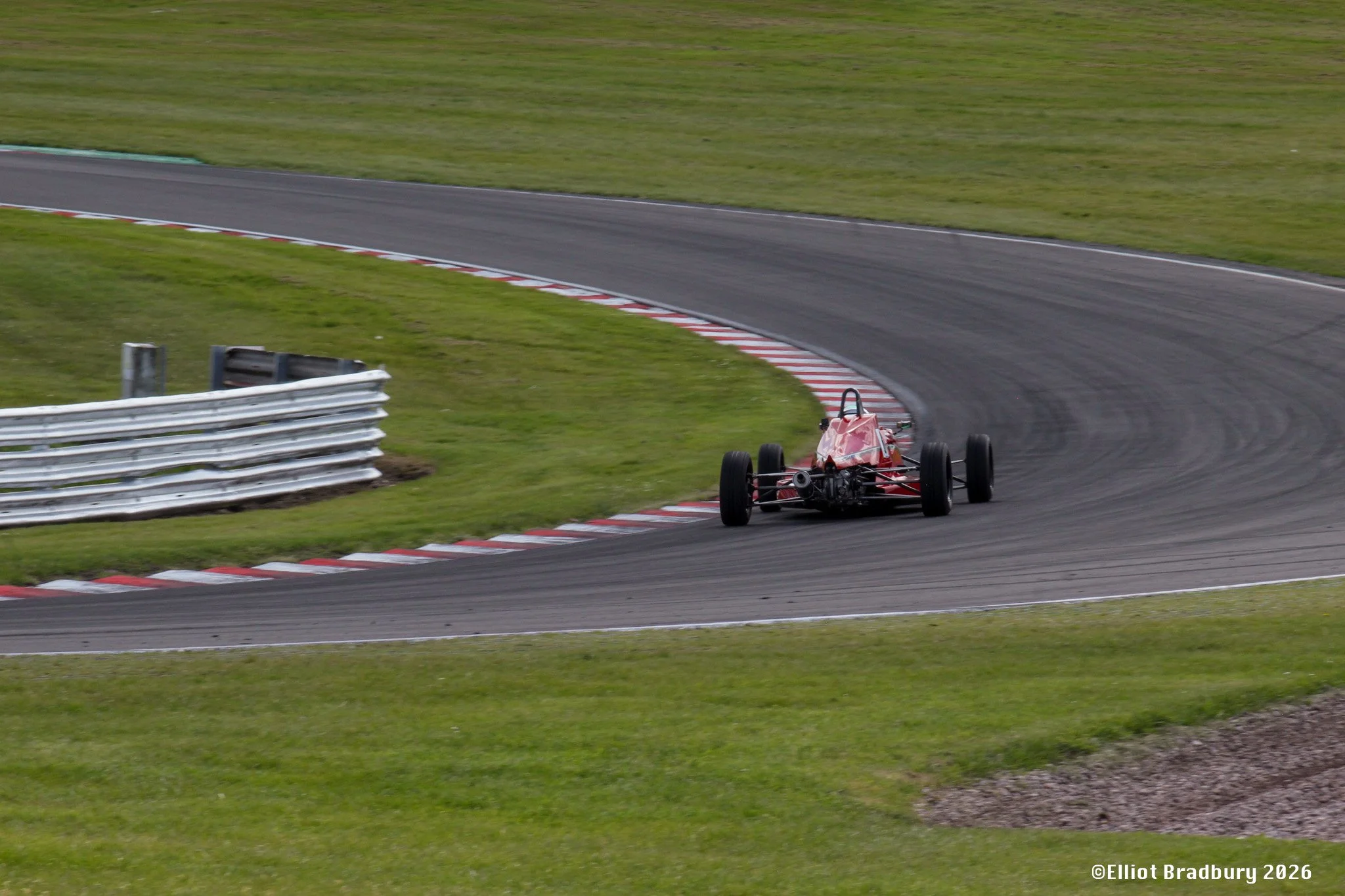 A red racing car on a race track taking a sharp right turn with grass on either side. The front of the car is angled into the turn while the rear wheels are slightly on the curbing. The track has red and white painted kerbs along the edge.