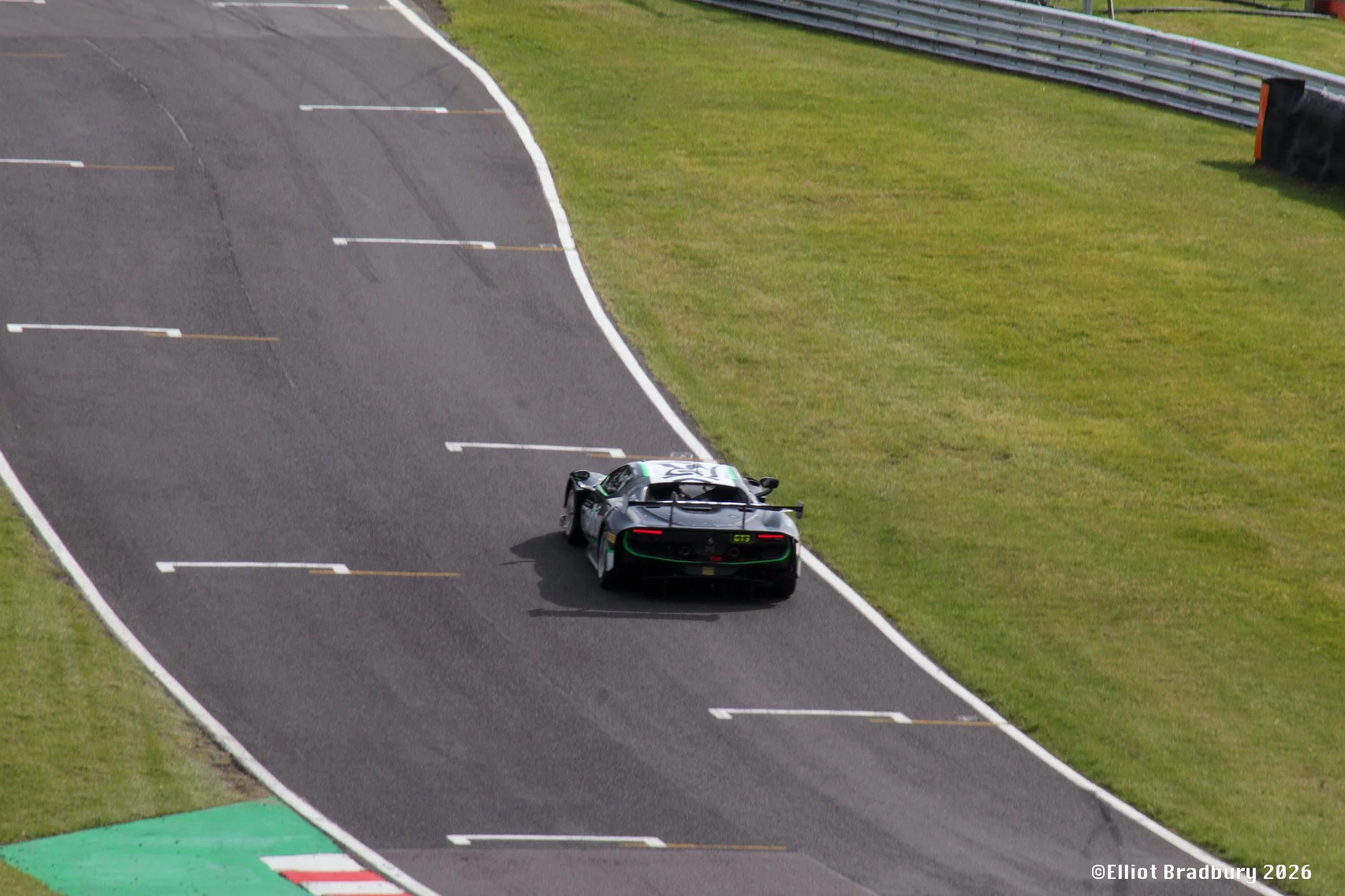 A black race car driven by a driver wearing a helmet on a race track, approaching a corner with a grass area on the right and safety barriers along the edge of the track.