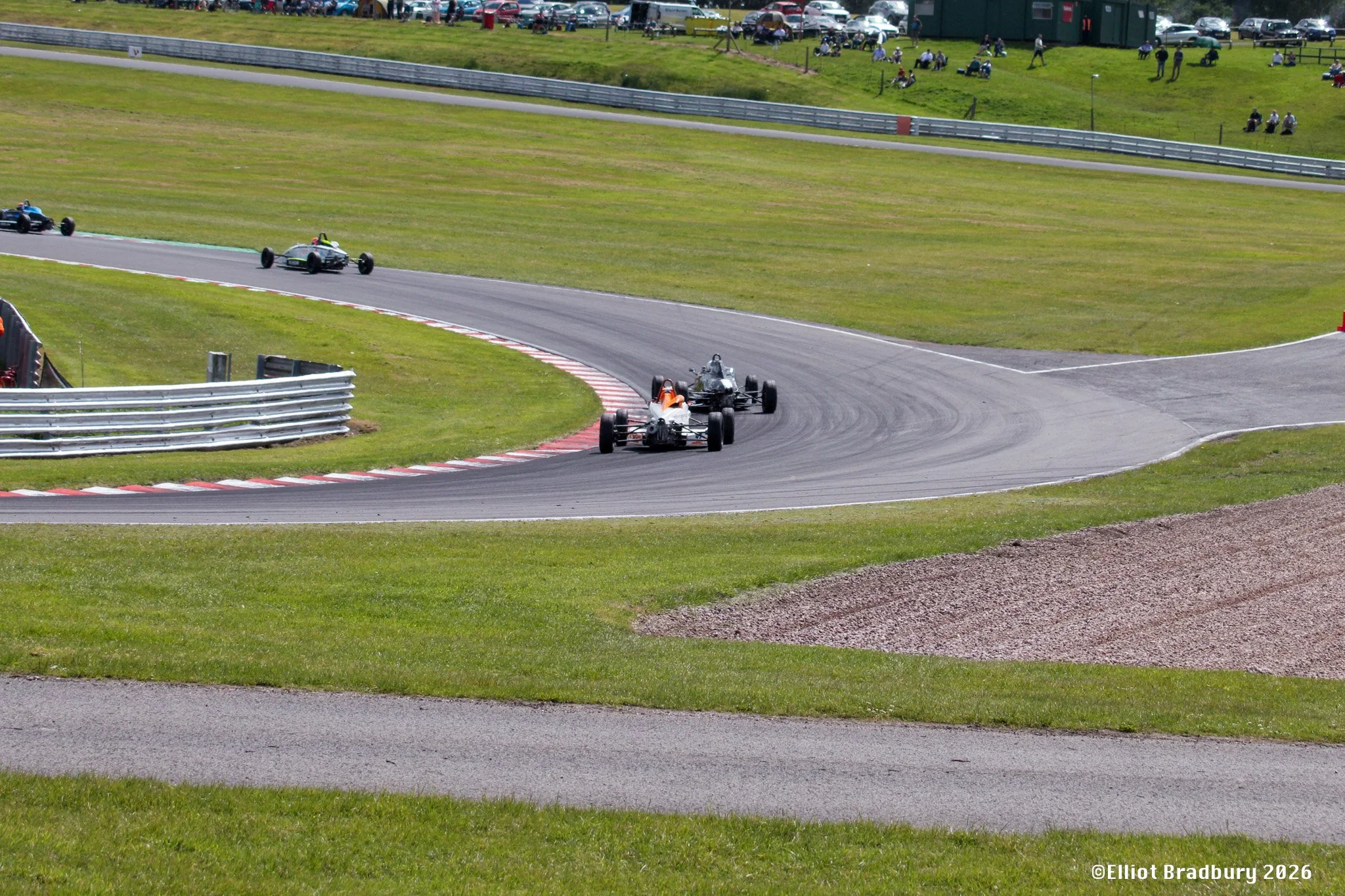 Race cars navigating a curve on a racetrack with grass and spectators in the background.