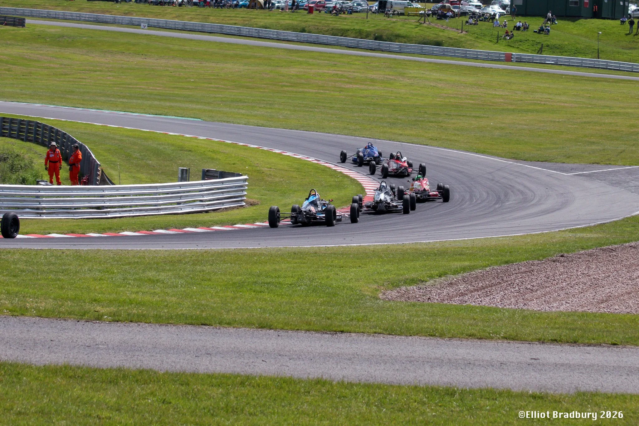 Race cars navigating a turn on a racing track, with two officials in red suits standing on the grassy area near the track and spectators in the background.