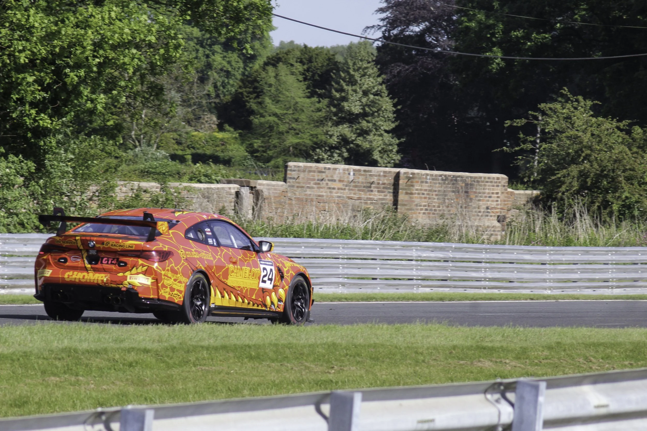 A brightly colored race car with a red and yellow dragon design and the number 24 on its side, driving on a road with green grass and trees in the background.