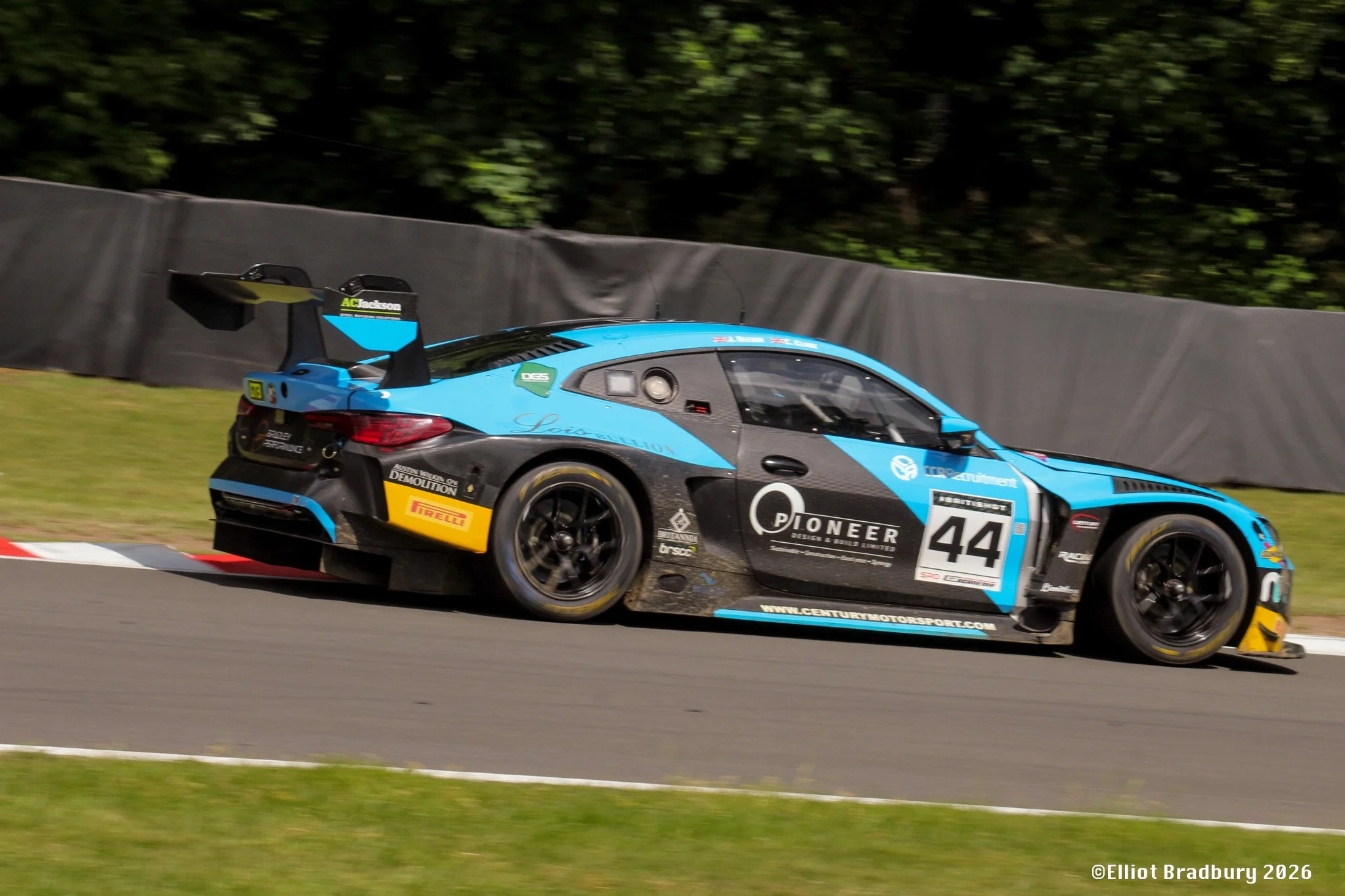 A race car on a track, painted in blue and black with various sponsor logos, racing past a dark gray barrier with green trees in the background.