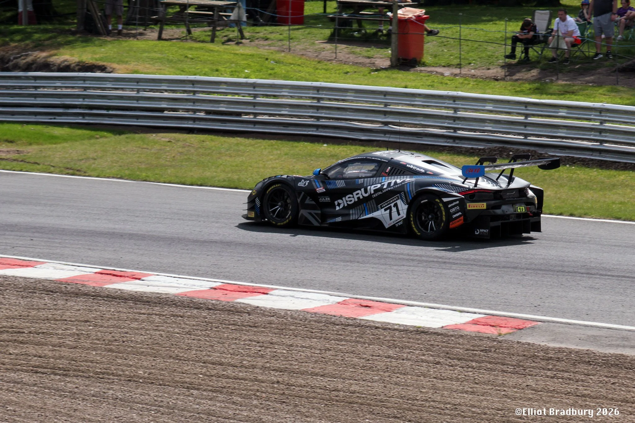 A black racing car on a race track, with spectators watching from a grassy hill behind a safety barrier.