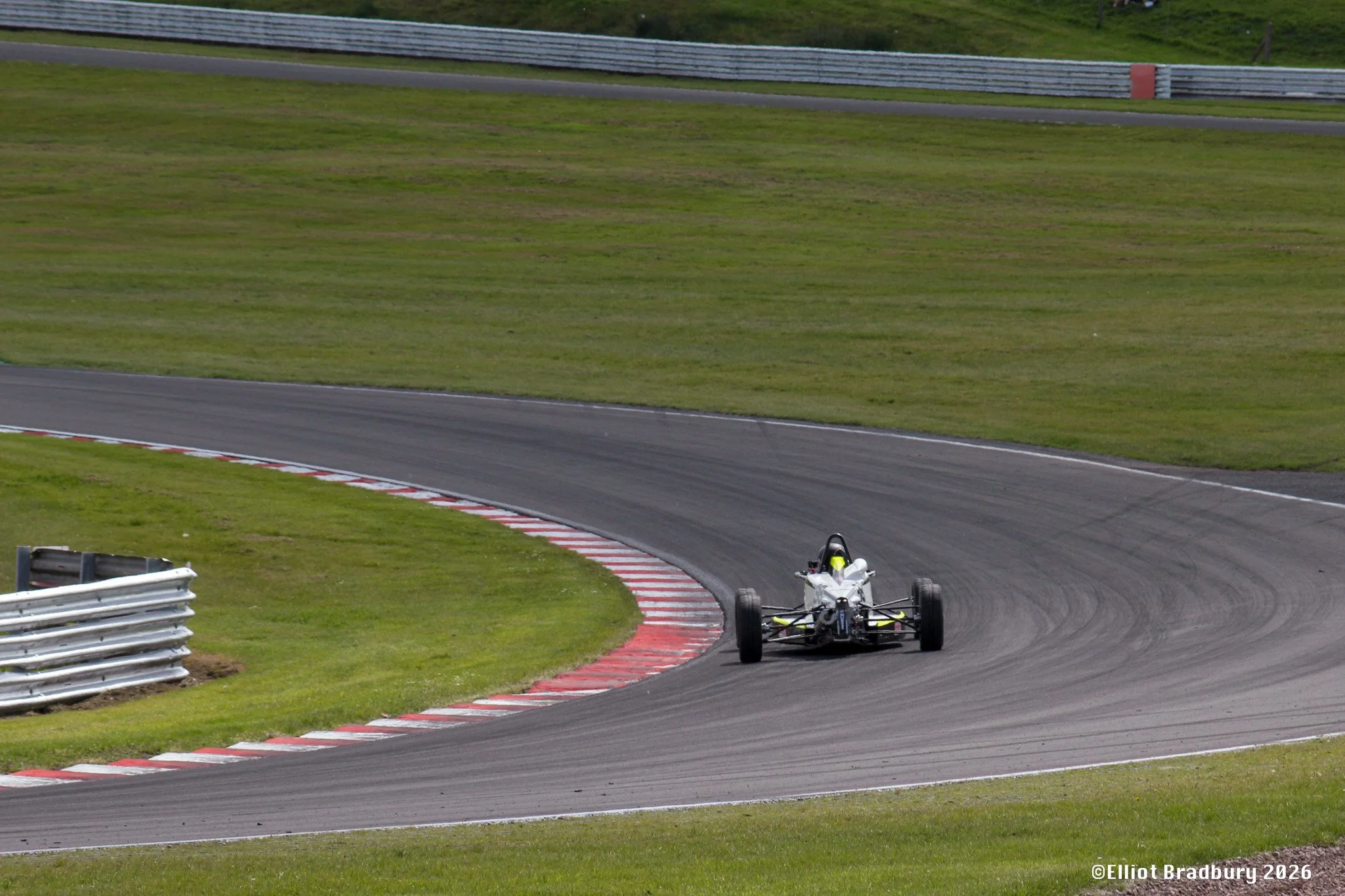 Race car on a curving race track surrounded by grassy areas with guardrails.