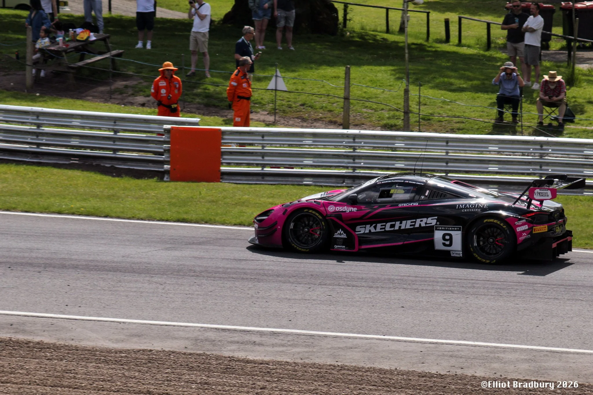 A pink and black race car driving on a race track with spectators watching from the grassy area behind a safety barrier. Two individuals in orange safety suits are standing near the fence, and several spectators are seated or standing, some with hats