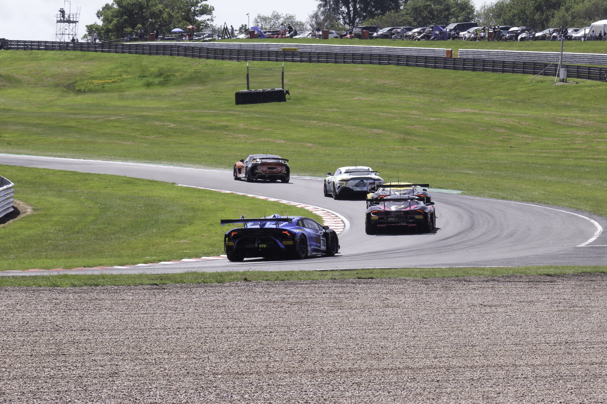 Multiple race cars driving on a curving racing track with green grass on either side and spectators in the background.