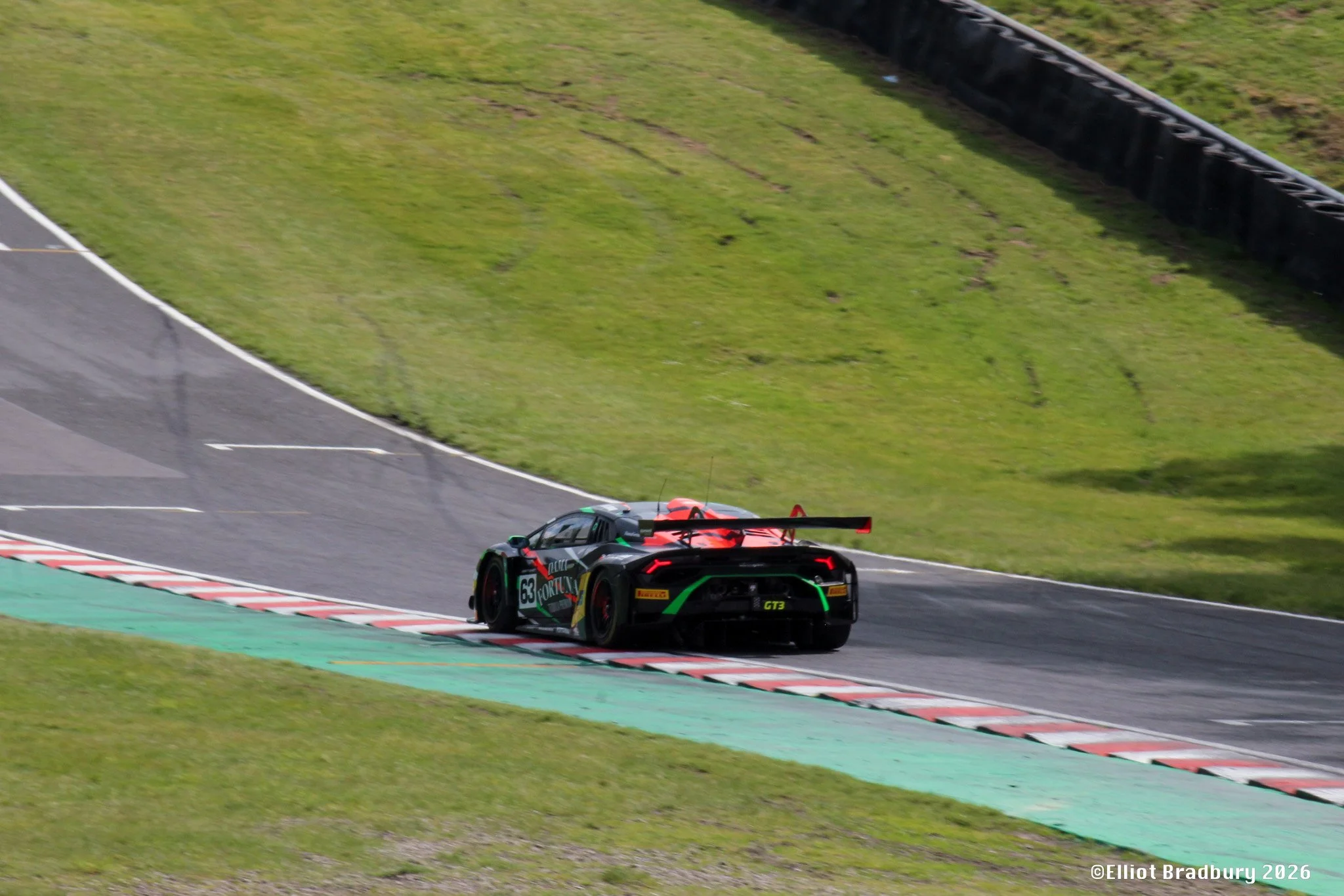 A black and green race car with the number 63 on its side speeding on a racetrack, following a curve with a grassy area and tires along the edge.