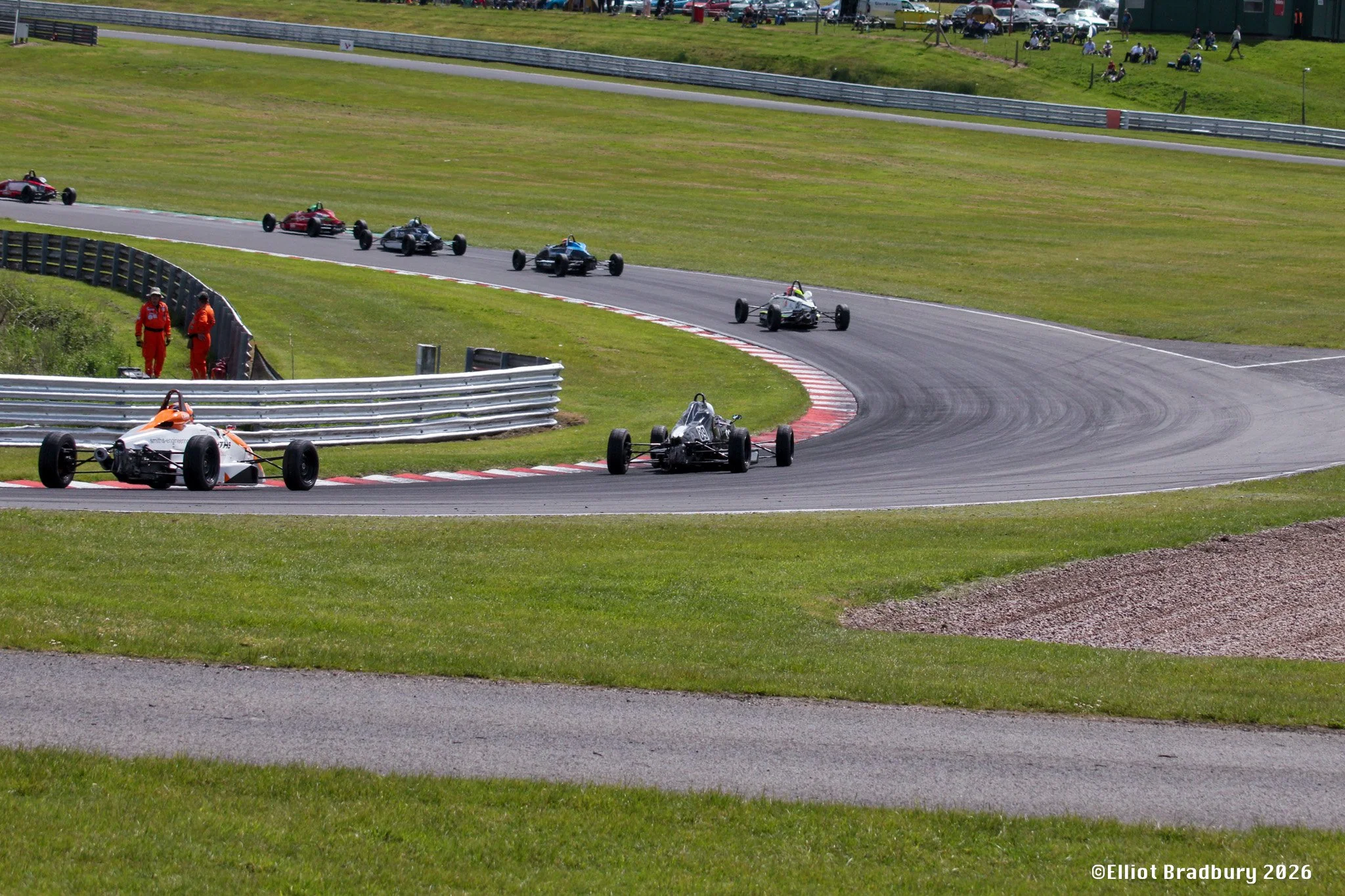 Several open-wheel race cars on a race track, with some cars approaching a turn and others following behind. Two people in red uniforms stand near the track's safety barrier. Spectators can be seen on the grassy hillside and in the background, with s