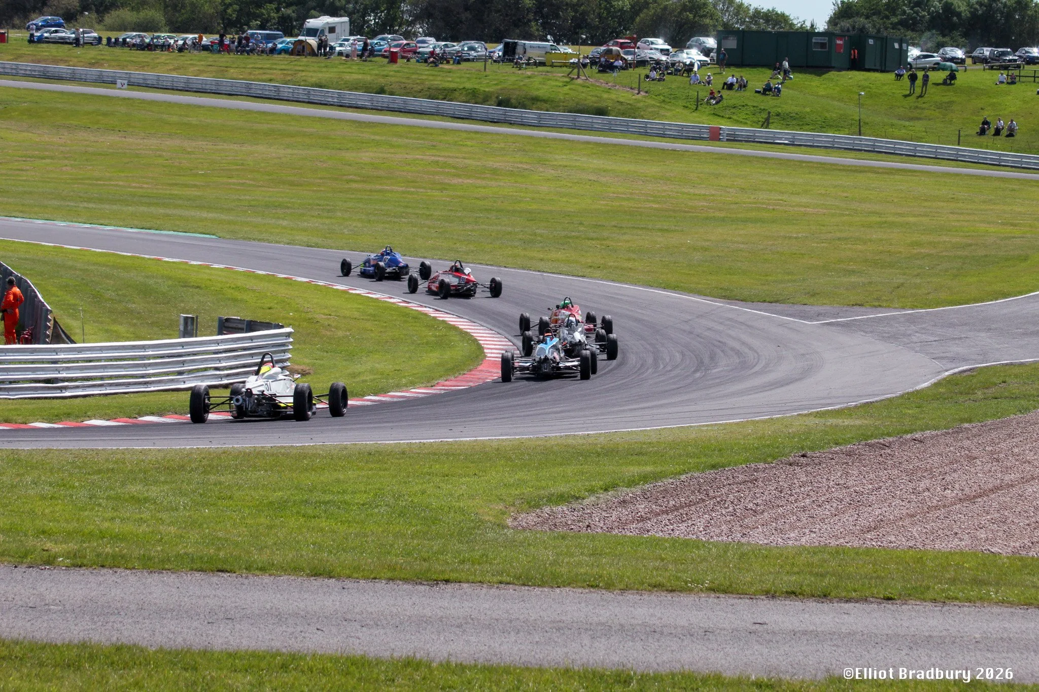Four open-wheel race cars on a racetrack during a racing event, with cars in the background and spectators sitting on the grassy hillside.