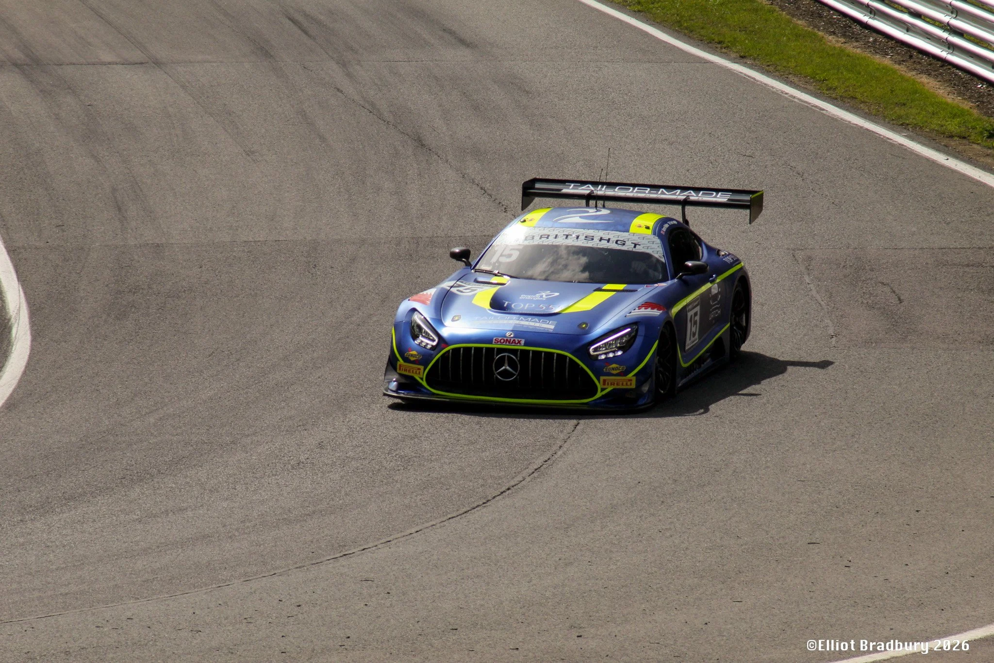 A blue race car with yellow accents, number 15, on a race track with tire marks, approaching a turn, with green grass and safety barriers on the side.