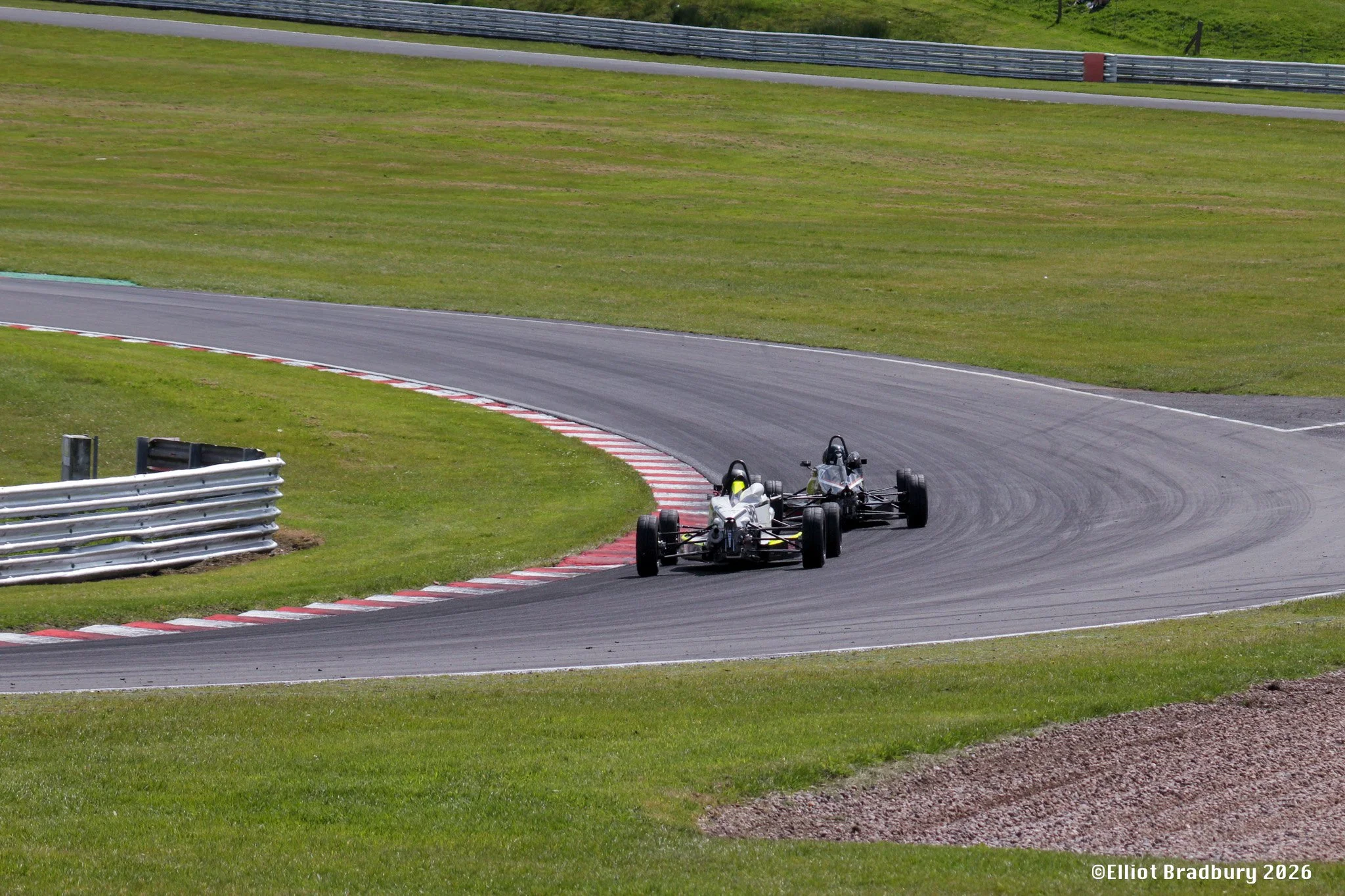 Two race cars on a curved section of a race track with green grass on either side and safety barriers along the track.