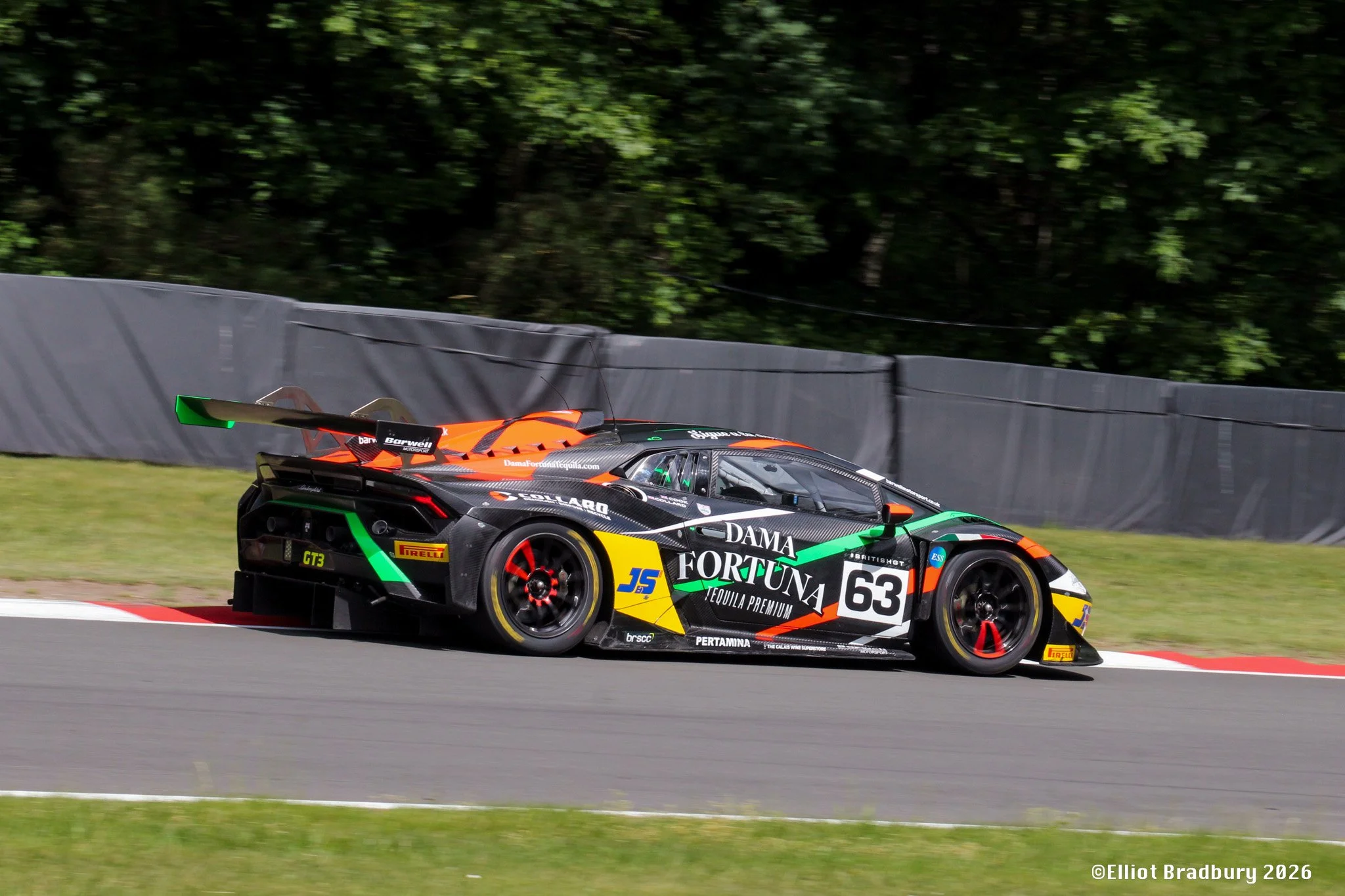 A race car on a track with a black, orange, and green paint job, displaying the number 63 and various sponsor logos, speeding past a grassy area and a black barrier with a wooded background.