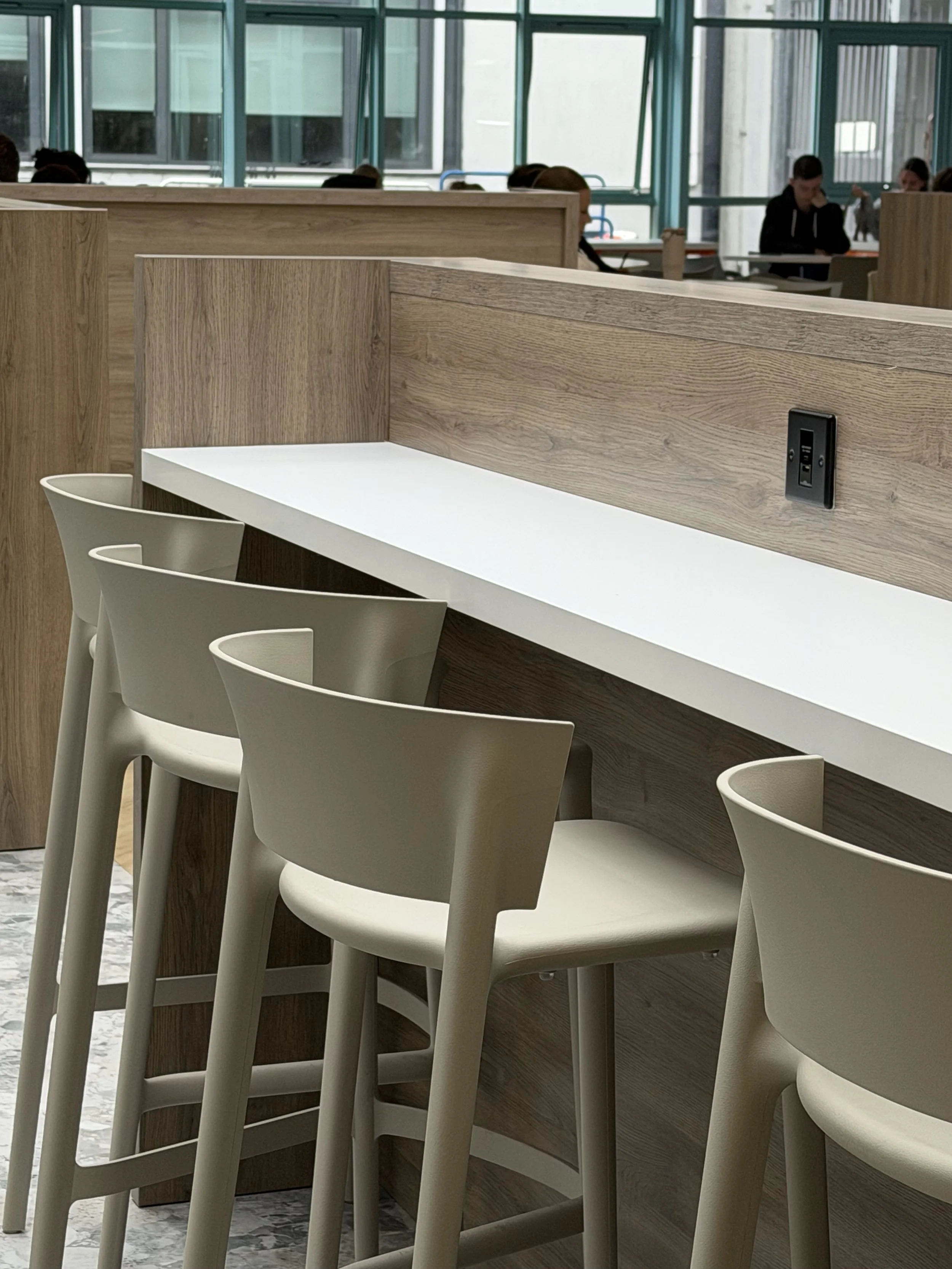 Interior of a modern cafe or study area with a white countertop and beige chairs, wooden dividers, and large windows with a view of buildings outside.