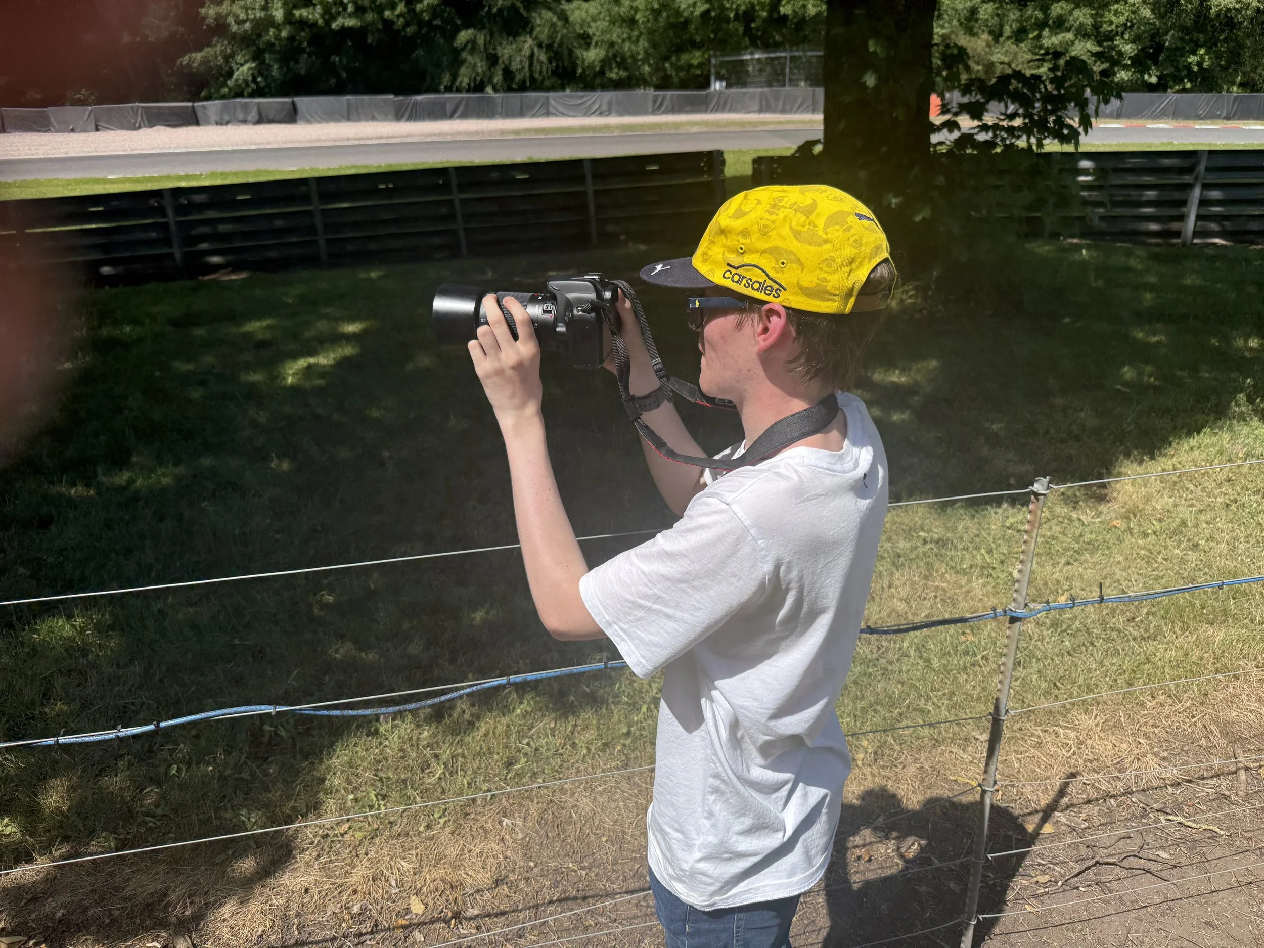 A young person wearing a yellow cap with black design and glasses is holding a camera and photographing a racetrack behind a wire fence.