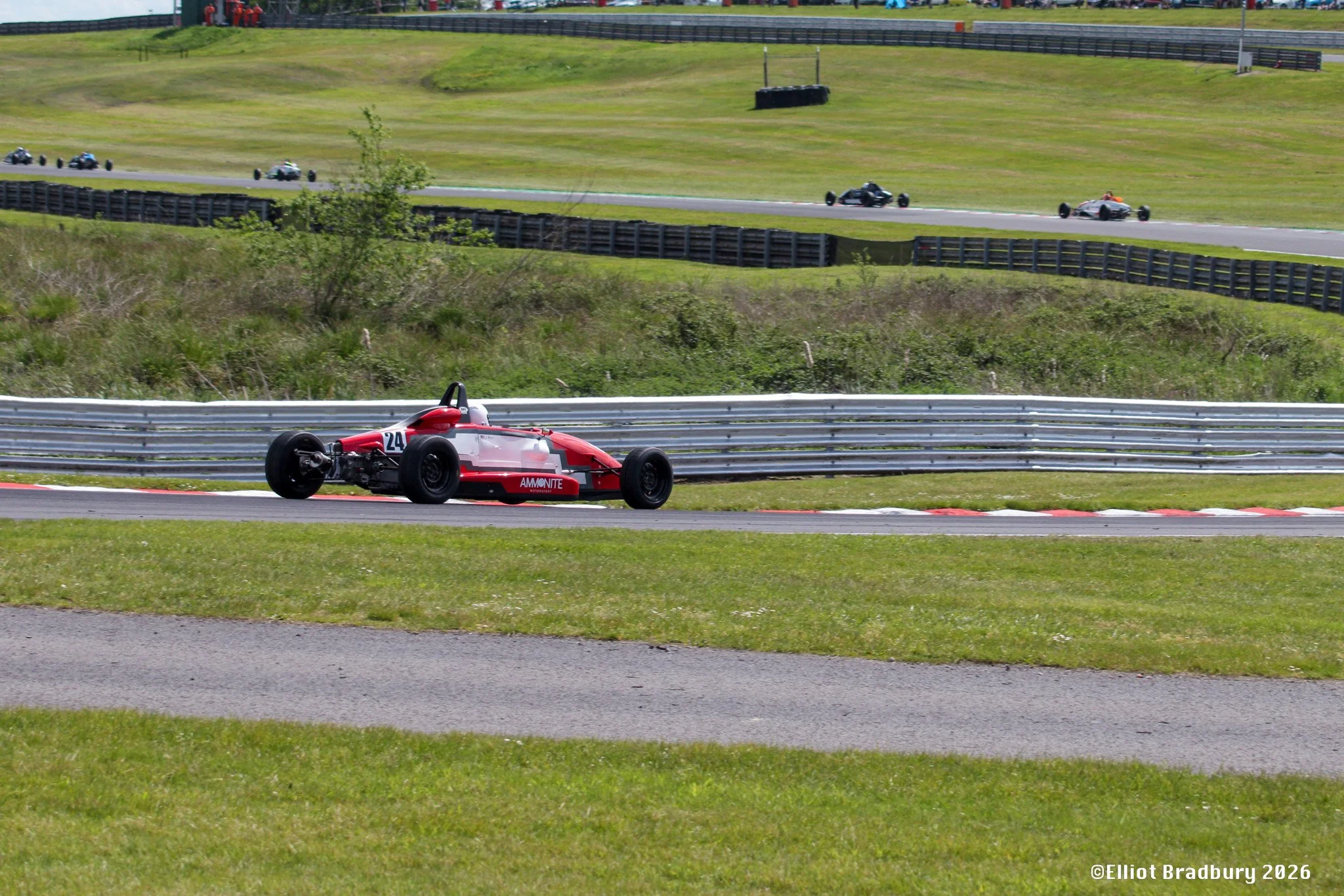 A red and white racing car with the number 24 on it going around a race track, with green grass, barriers, and other race cars in the background.